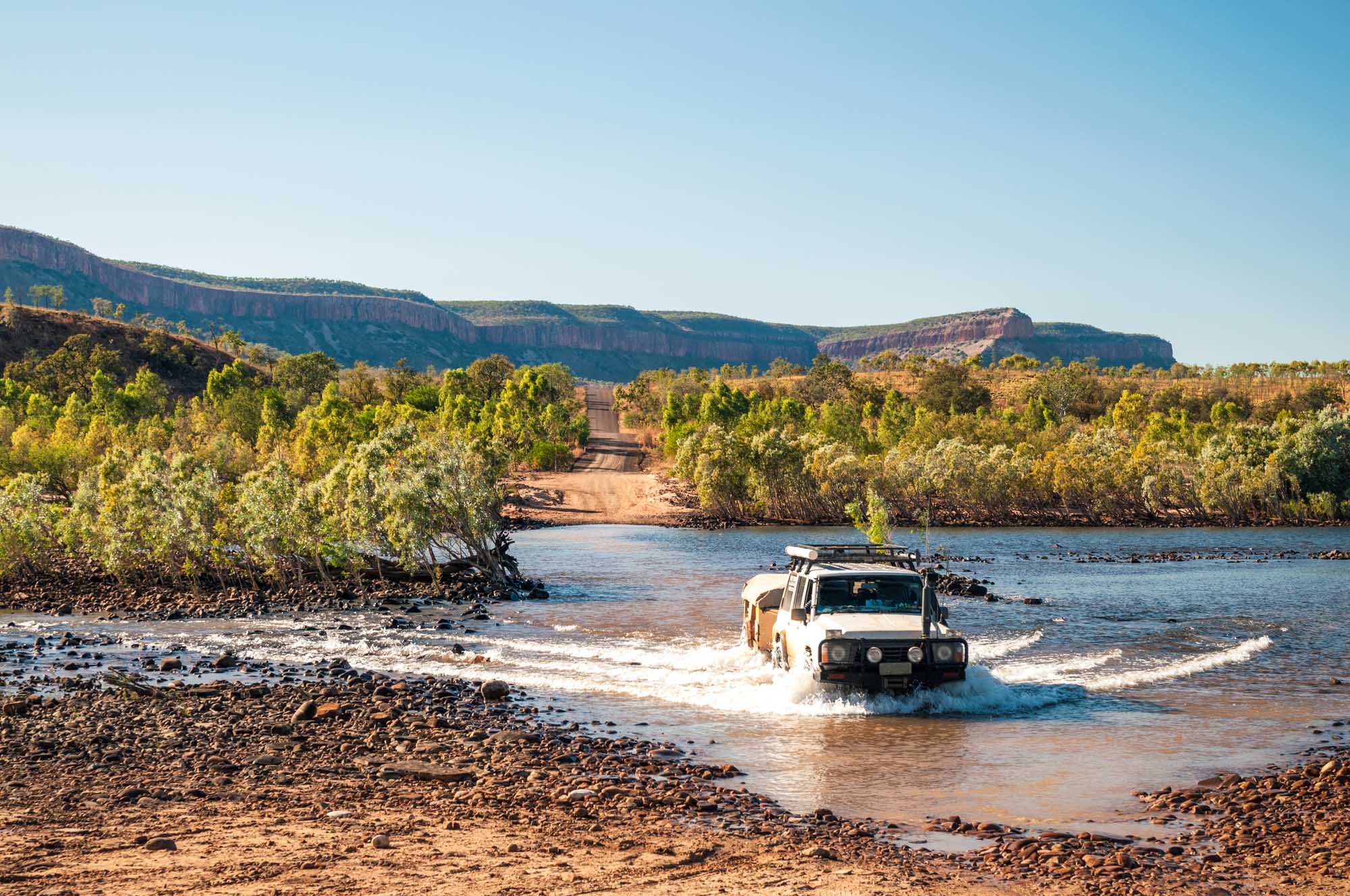 A white four-wheel-drive vehicle equipped with a roof rack crosses a wide, shallow river during an off-road expedition in The Kimberley region of Western Australia. The landscape features dramatic red-rock plateaus, dense mangrove vegetation, and clear blue skies, showcasing the rugged beauty of this remote Australian destination.