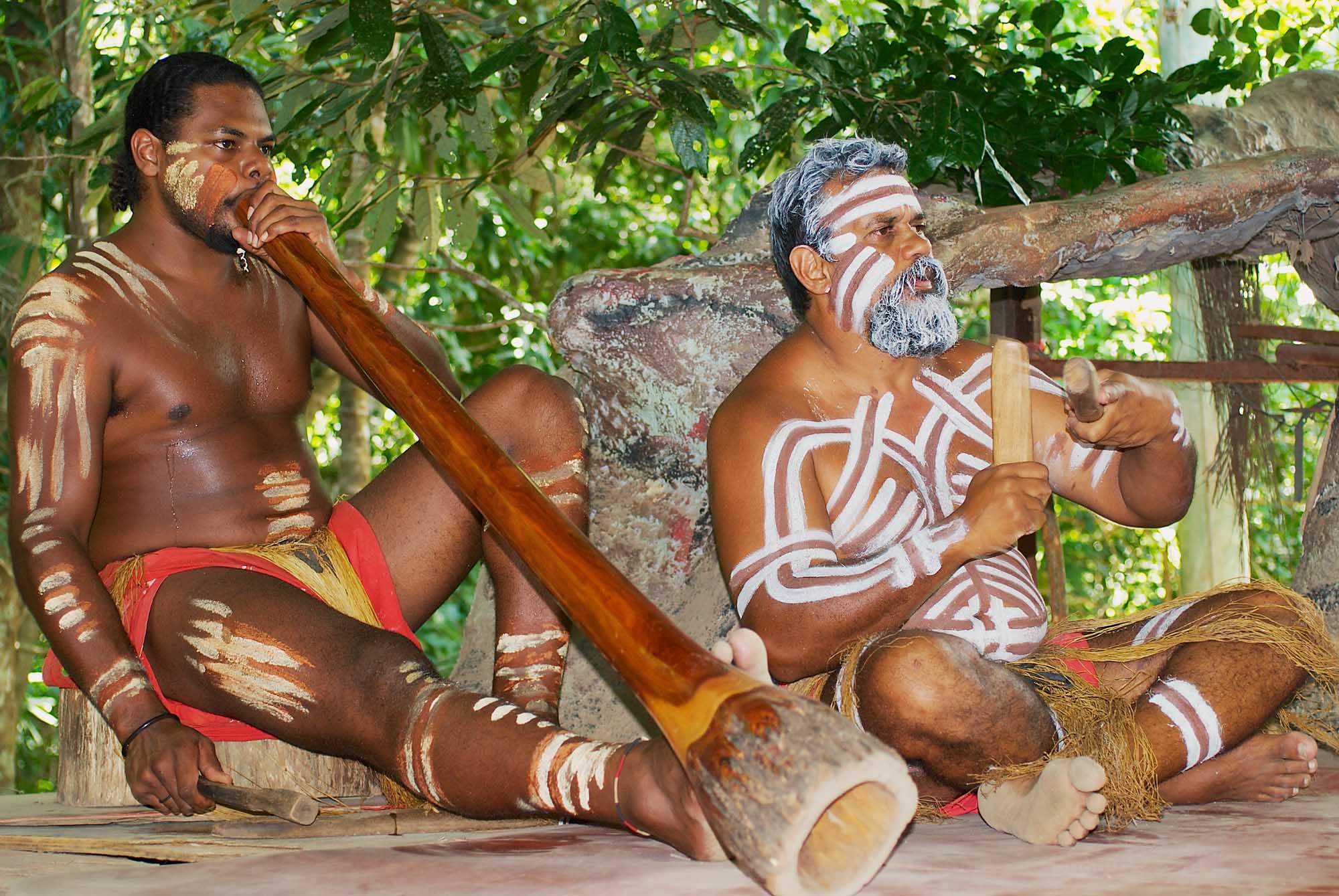 Two Aboriginal performers demonstrate traditional music and instrumentation at the Tjapukai Culture Park in Kuranda, Australia. The men display traditional body paint and decorations while playing indigenous instruments in a natural rainforest setting.