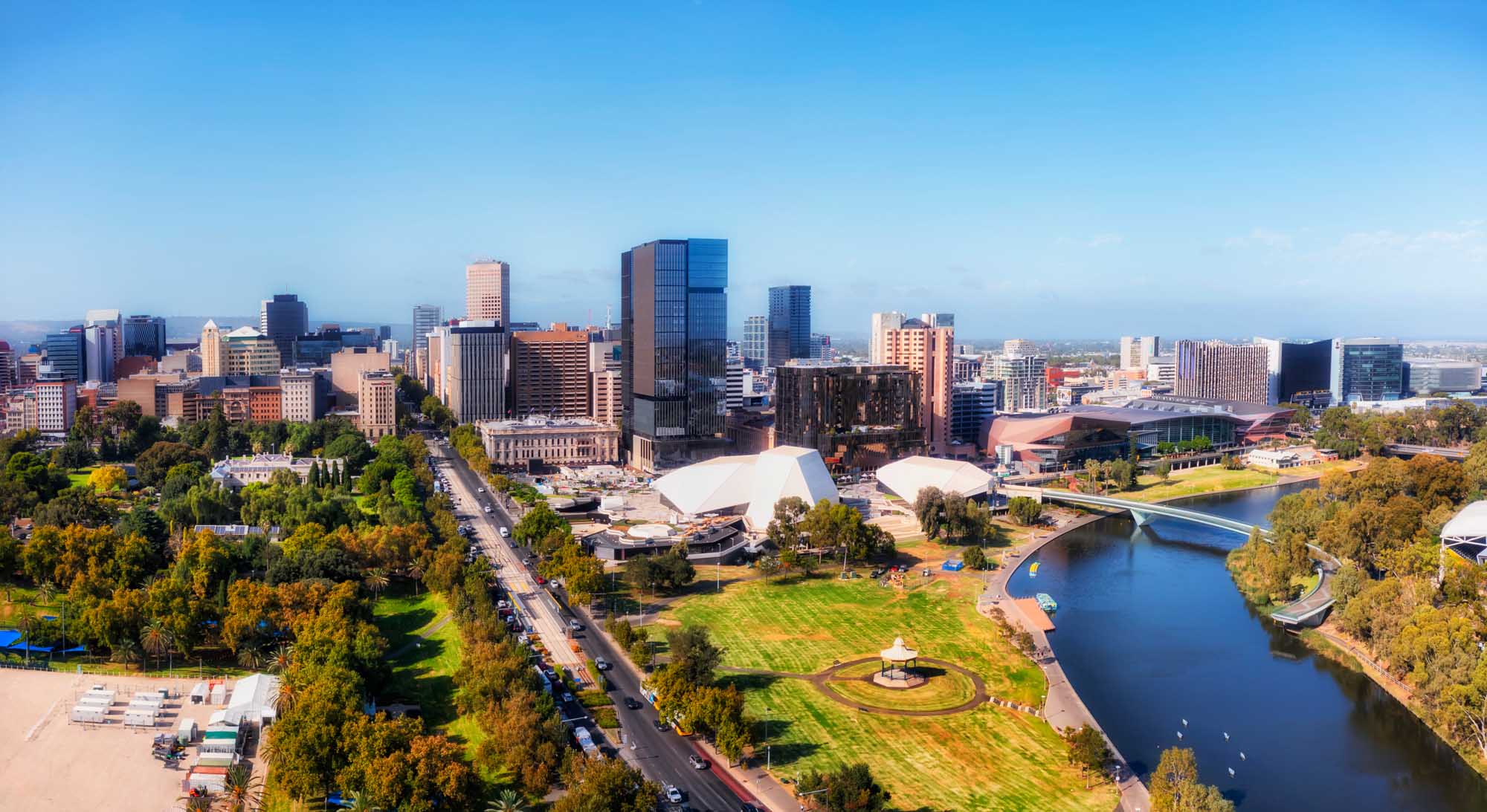 An aerial photograph of Adelaide's central business district skyline along the Torrens River in South Australia. The image showcases modern high-rise buildings, cultural institutions with distinctive white architectural features, manicured parklands, and the river's curved path through the urban landscape under clear blue skies.