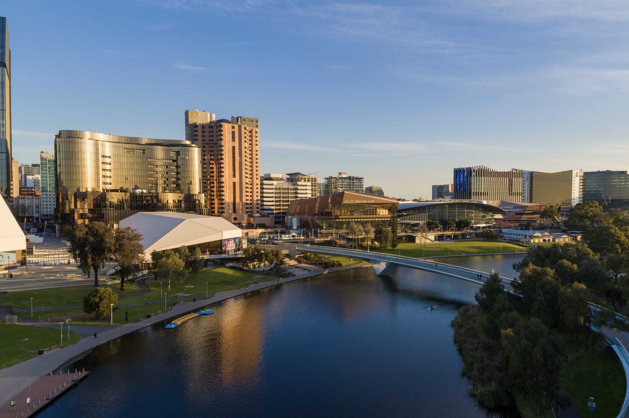 A vibrant riverfront cityscape in Adelaide, South Australia, featuring modern cultural institutions including theaters and museums along the Torrens River, with contemporary glass and copper-clad buildings illuminated by golden hour light.