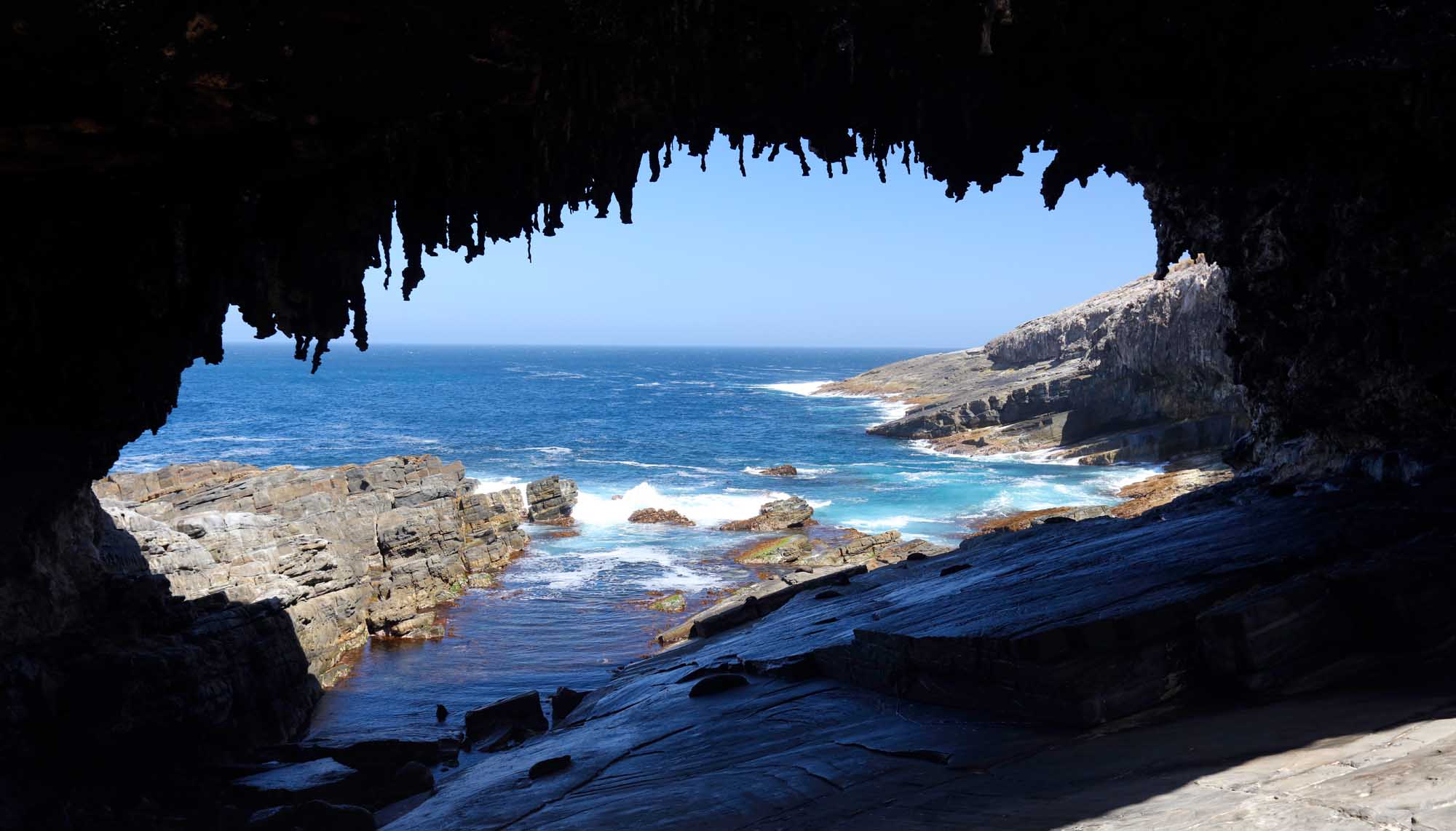 A dramatic seascape framed through a natural sea cave opening at Admirals Arch on Kangaroo Island, revealing turquoise ocean waters, rocky outcrops, and rugged coastline under clear blue skies. The photograph captures the interplay of shadow and light as the cave frames the pristine coastal landscape beyond.