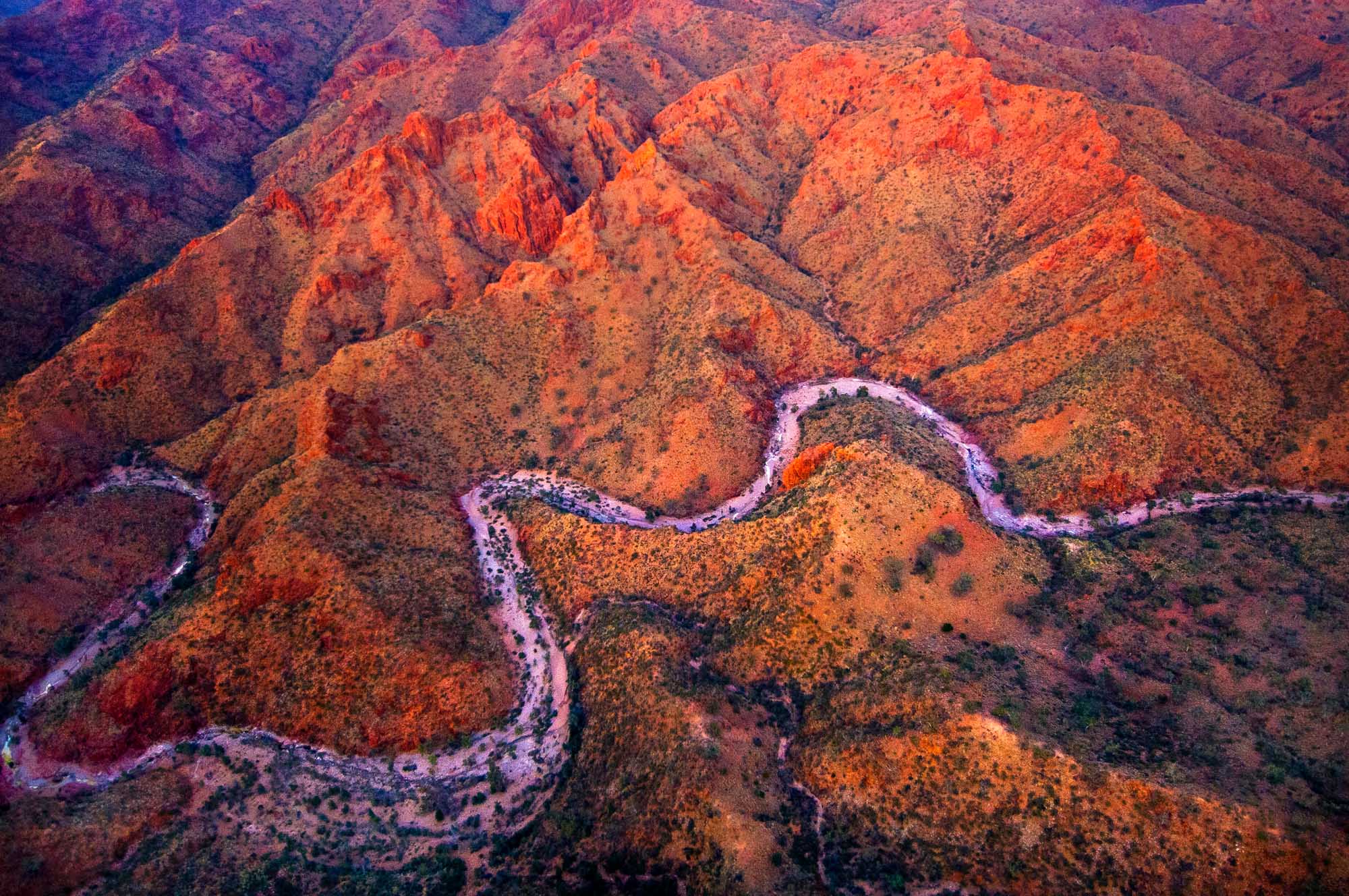 A stunning aerial photograph taken at first light over the town of Arkaroola in South Australia's Flinders Ranges, captured from a helicopter. The landscape displays dramatic ridges and valleys with rich orange, red, and purple hues typical of the ancient mountain range, with a winding river cutting through the terrain below.
