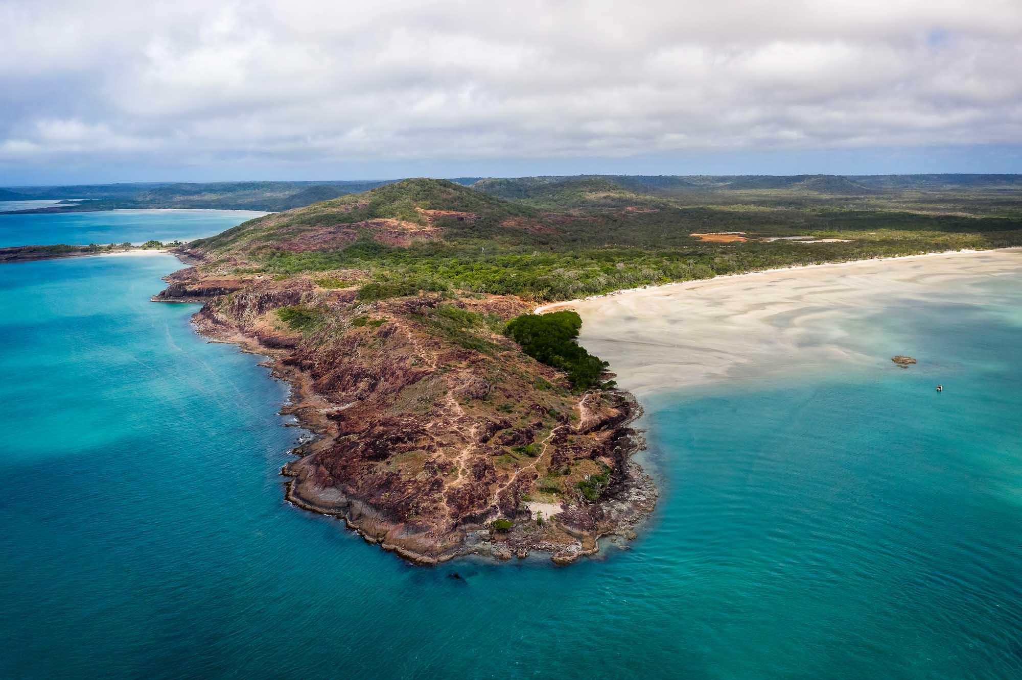 An aerial photograph of the rugged tip of Cape York Peninsula in tropical northern Australia, featuring distinctive red and purple volcanic rock formations, lush green vegetation, pristine white sand beaches, and turquoise waters. The dramatic rocky headland extends into the deep blue ocean, showcasing the region's unique geological character and pristine natural landscape.