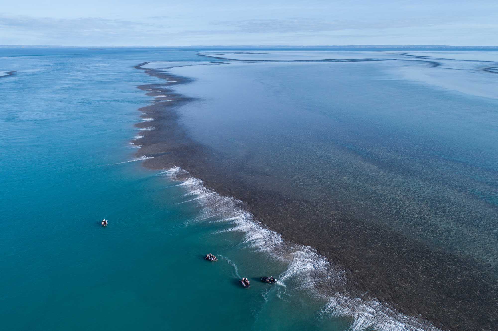 An aerial perspective of a dramatic coastal landscape featuring a distinctive dark reef or sandbank formation with white wave breaks along its edges, surrounded by brilliant turquoise waters. Several small boats are visible in the shallow turquoise waters, suggesting this is a popular tourist or expedition destination.
