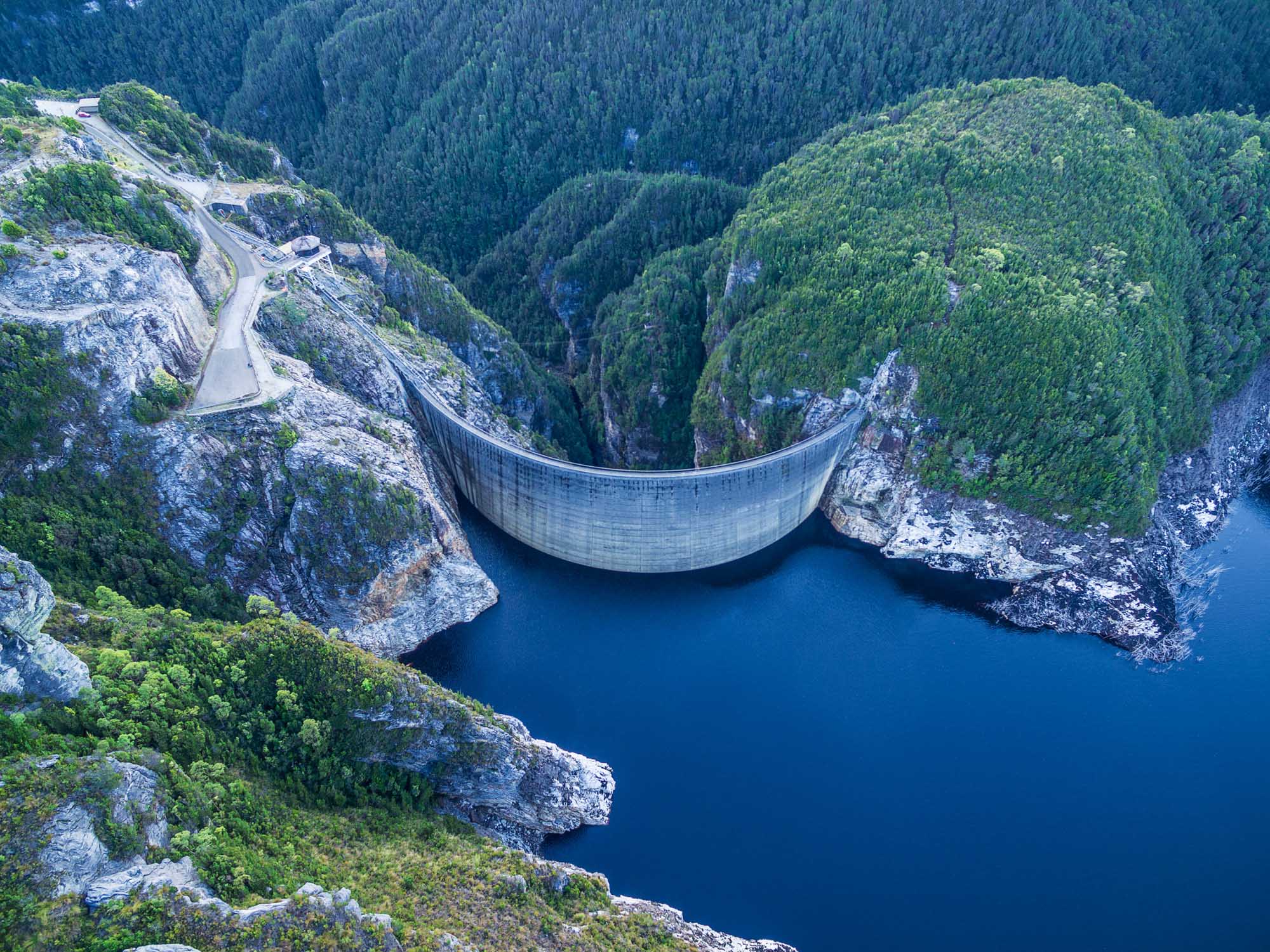 An aerial photograph of Gordon Dam, a large curved concrete dam structure spanning across a deep forested valley with brilliant blue water behind it. The dam is situated in Southwest Tasmania, surrounded by dense coniferous forest and rocky cliff faces, with a winding road visible on the left side leading to the dam.