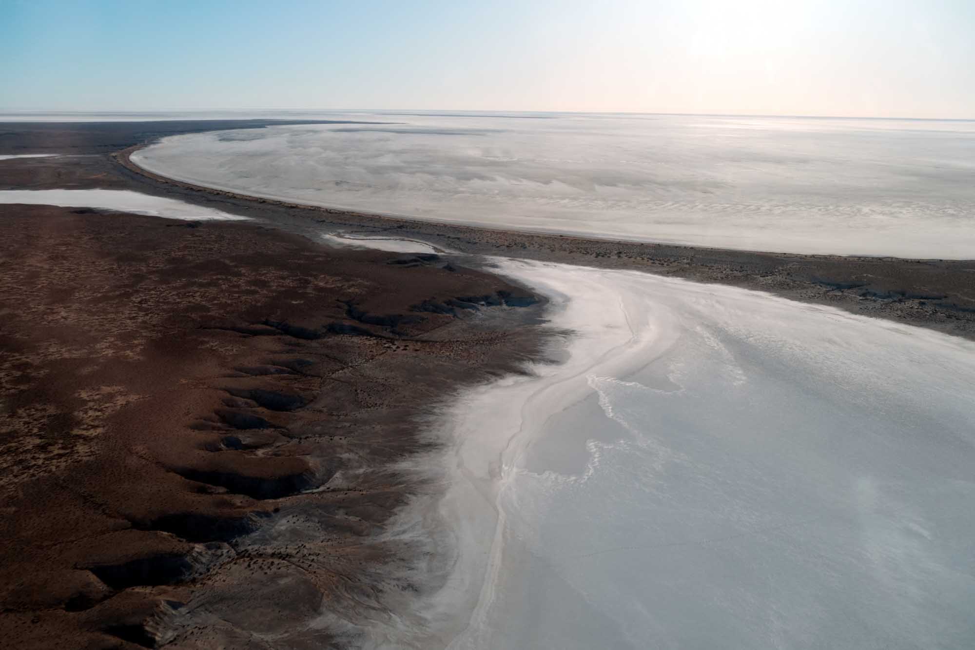 An aerial photograph of Kati Thanda–Lake Eyre, a vast endorheic salt lake basin in South Australia's Far North region. The image captures the striking contrast between the dark, mineral-rich shoreline and the pale, crystalline salt flats, with winding water channels cutting through the landscape.