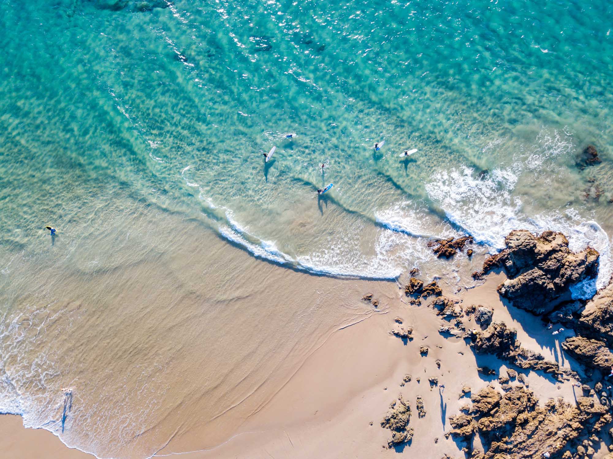 An overhead drone photograph of The Pass at Byron Bay, Australia, showing multiple surfers in the turquoise water with breaking waves, golden sand, and distinctive rocky outcrops along the shoreline on a clear sunny day.