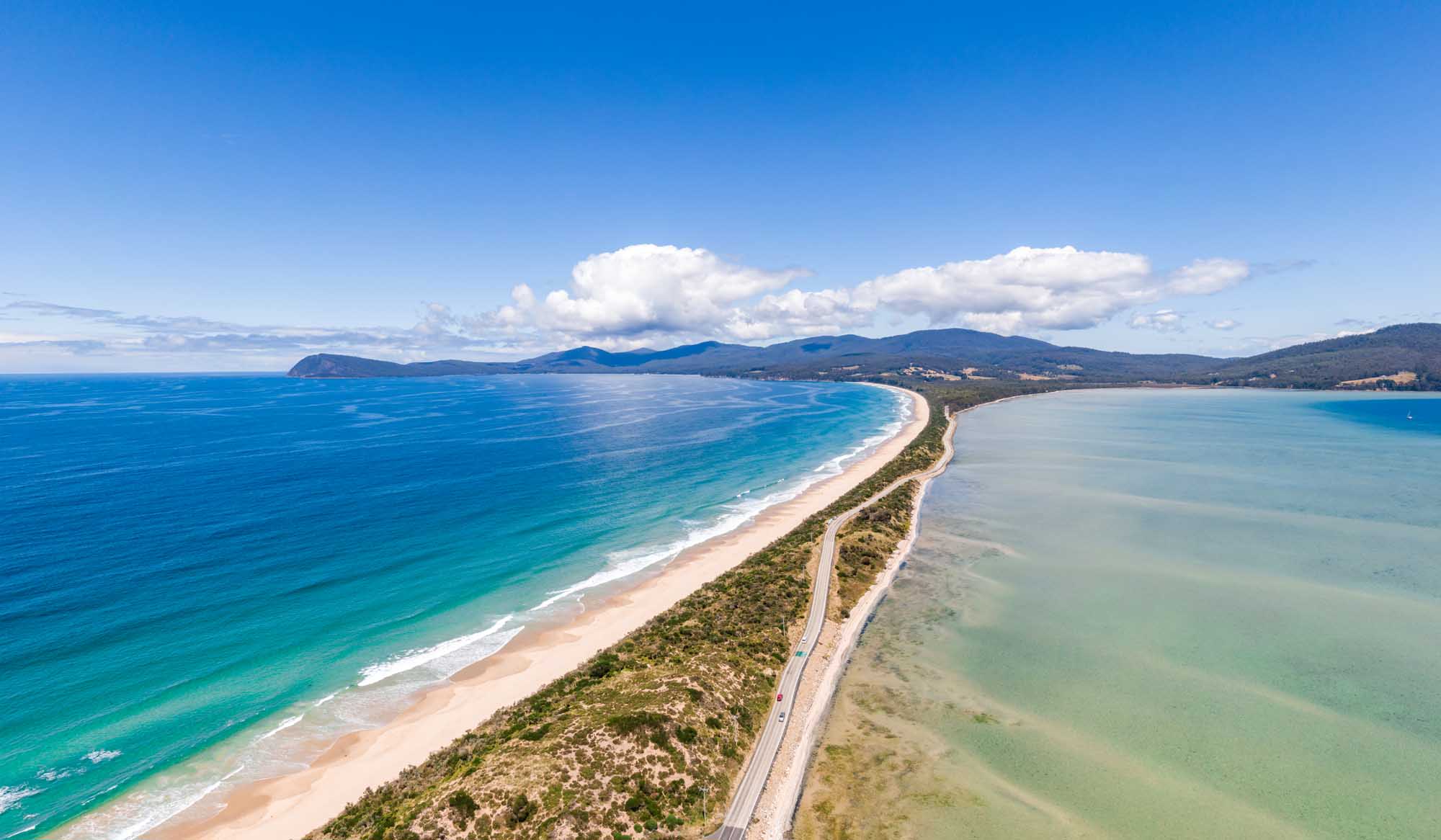 A stunning aerial drone photograph of The Neck, a narrow isthmus connecting North and South Bruny Island in southern Tasmania, Australia. The image showcases the dramatic contrast between the turquoise ocean waters on the left, the sandy beach, pristine green lagoon on the right, and forested hills beyond, with mountains visible in the distance under a partly cloudy sky.