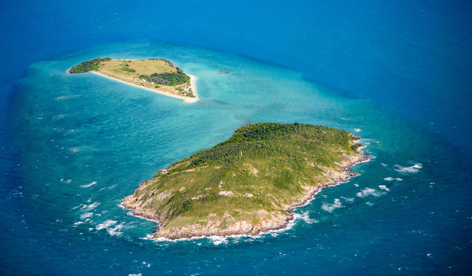 An aerial photograph of two lush, forested islands surrounded by pristine turquoise waters in the Torres Strait. The larger island in the foreground features dense tropical vegetation covering rocky terrain, while a smaller sandy island is visible in the background, both framed by vivid blue ocean waters.