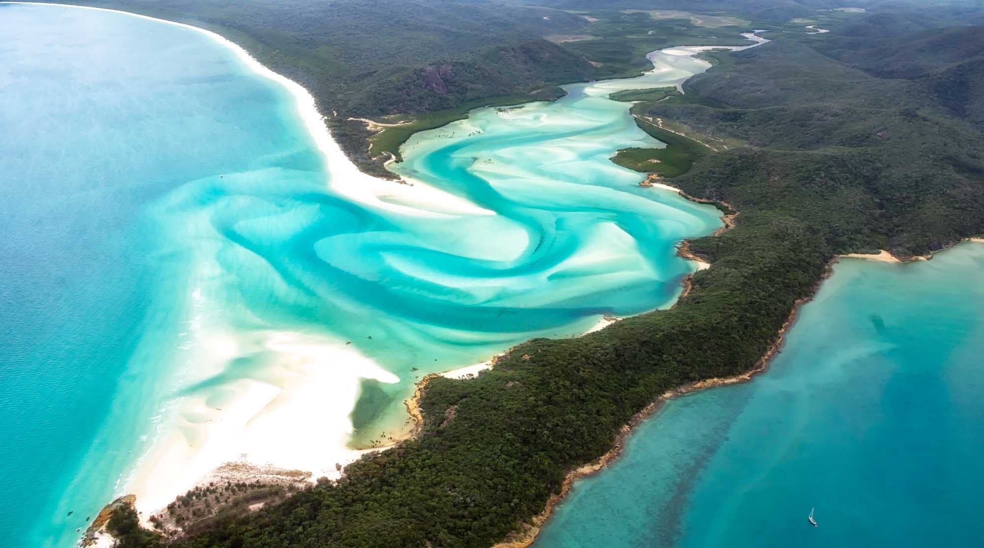 A stunning aerial photograph showcasing the Whitsunday Islands in Australia, featuring pristine turquoise waters, brilliant white sandy beaches, and lush green forests. The image captures the dramatic contrast between the vibrant aquamarine lagoons with their distinctive sandbars and the dense tropical vegetation covering the islands.