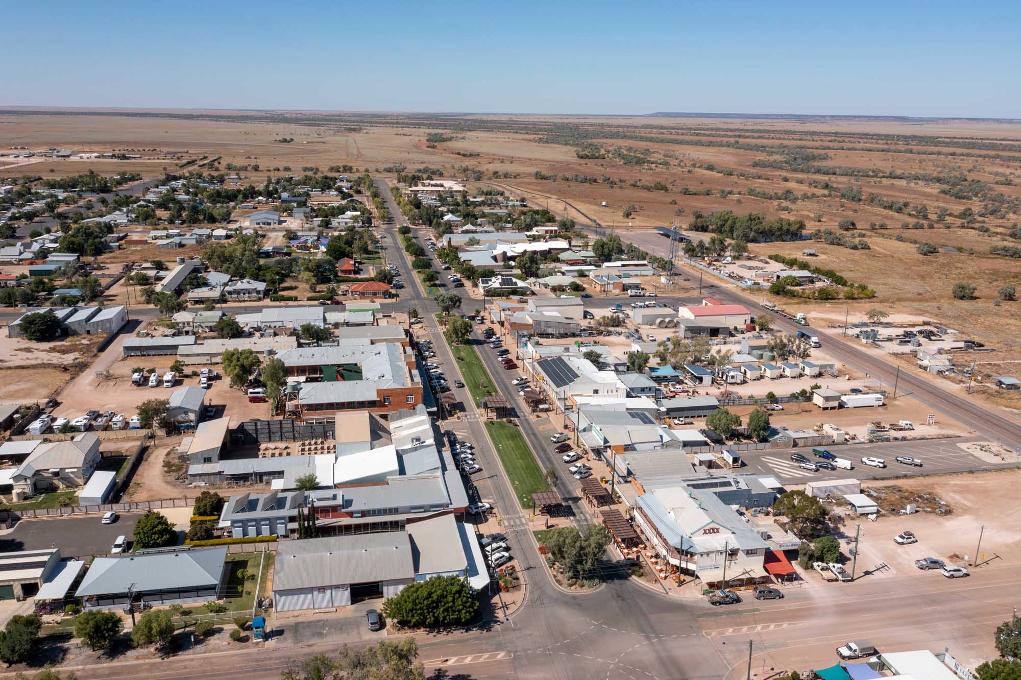 An aerial photograph of Winton, a rural town in central Queensland, Australia, showing the main commercial street lined with businesses, residential areas with scattered trees, and vast surrounding agricultural land under clear blue skies.