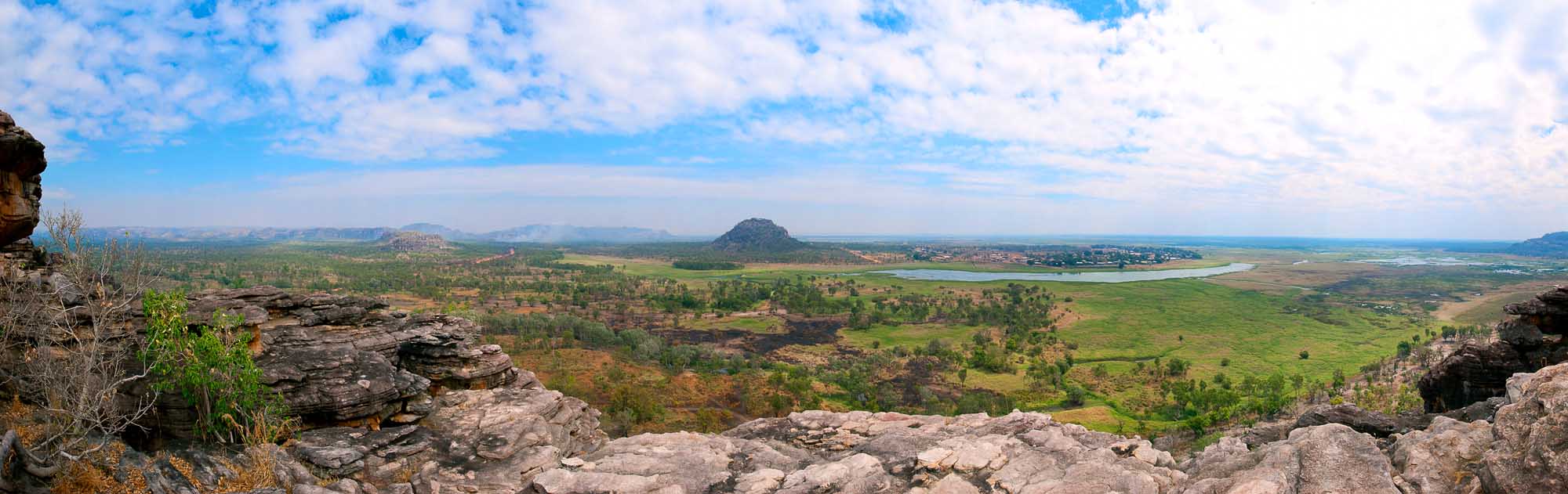 A sweeping panoramic view of Arnhem Land in Australia's Northern Territory, showcasing lush green valleys, scattered vegetation, distinctive rock formations in the foreground, and distant plateaus under a partly cloudy sky. The landscape features a mix of tropical woodlands, agricultural areas, and natural water features characteristic of this remote Australian region.