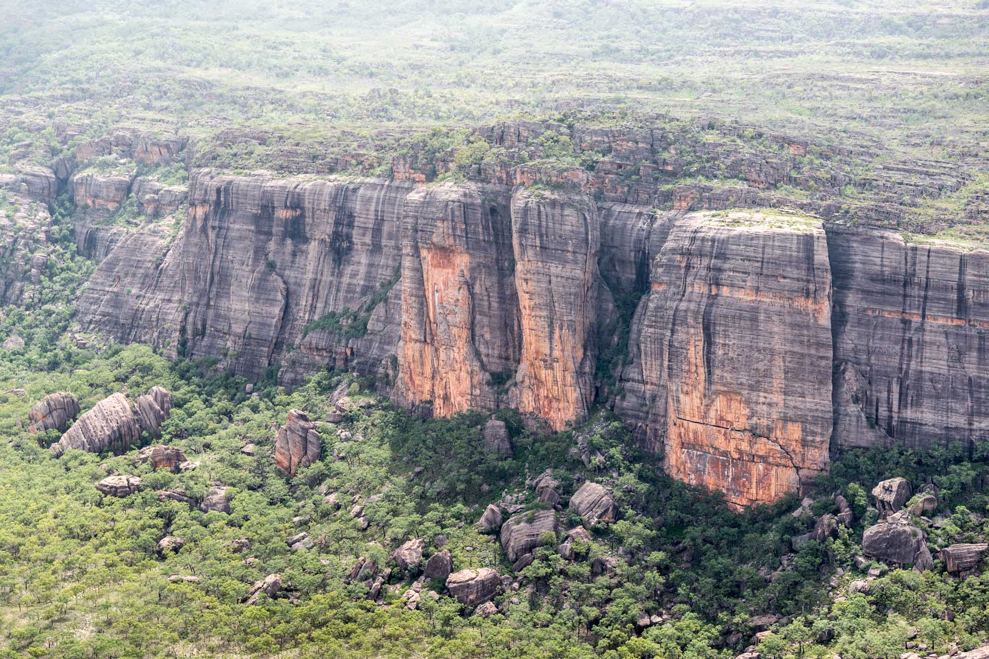 An aerial photograph of dramatic sandstone rock formations and plateau cliffs rising above lush green bushland in Australia's Northern Territory. The distinctive columnar basalt formations and layered red rock faces are characteristic of the rugged Arnhem Land escarpment, surrounded by sparse eucalyptus woodland.