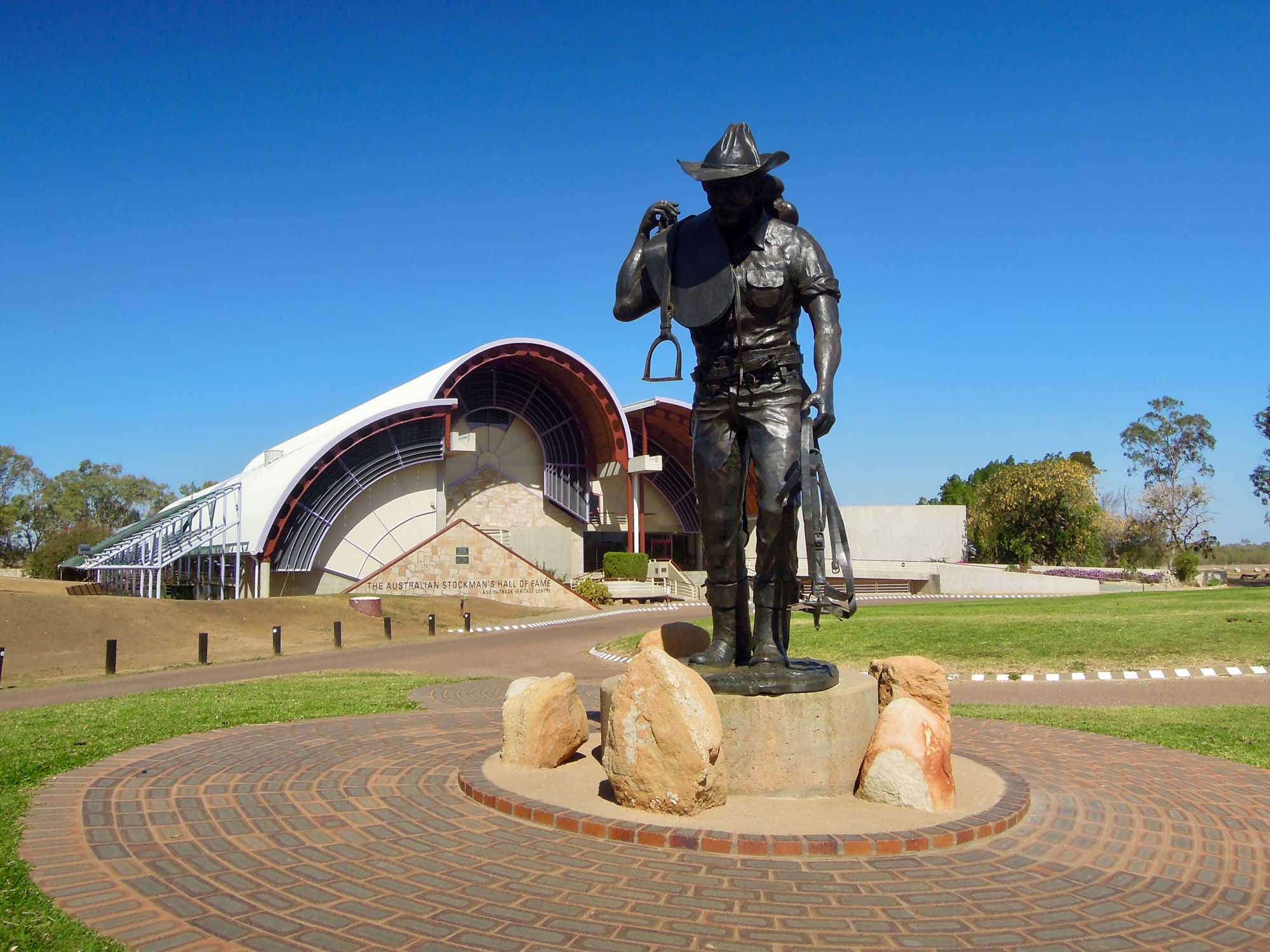 A bronze statue of a cowboy in traditional Australian stockman attire stands in the foreground holding a saddle and stirrup, positioned in front of the distinctive curved architecture of the Australian Stockman Hall of Fame building in Longreach, Queensland. The modern facility features a striking red and white curved roof design set against a clear blue sky.