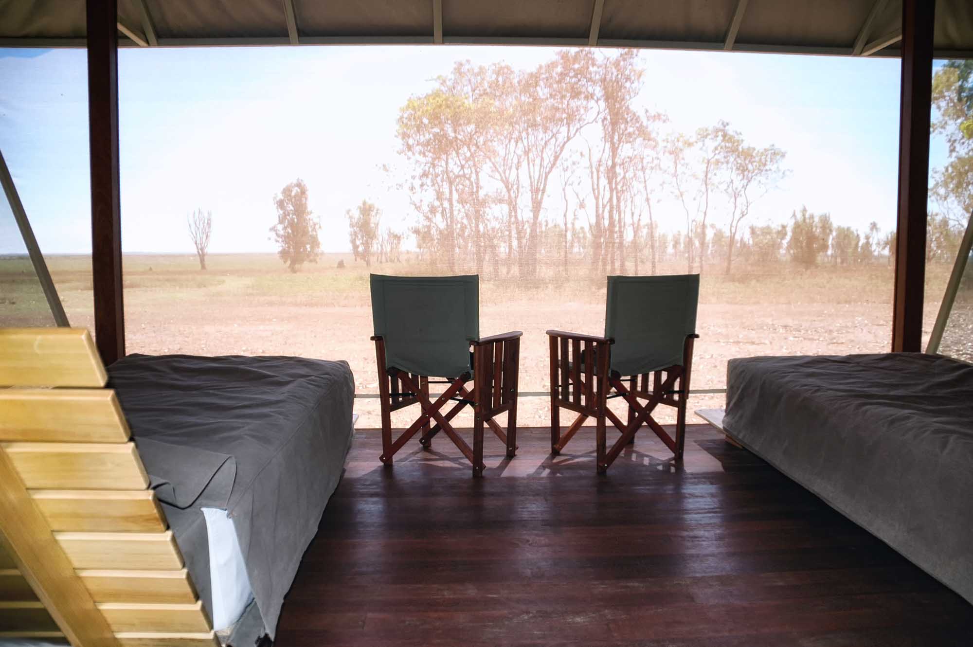 A spacious bedroom at Bamurru Plains Lodge in Northern Territory, Australia, featuring two beds with dark bedding and two director's chairs positioned on a wooden deck overlooking vast savanna plains dotted with sparse eucalyptus trees under a clear sky.