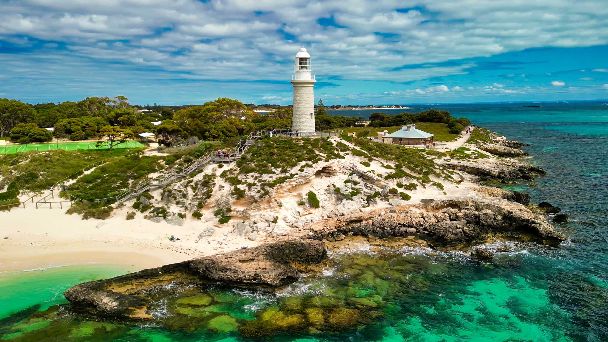 Aerial view of the iconic white Bathurst Lighthouse perched on a rocky headland at Rottnest Island, Australia. The scene captures pristine turquoise waters, sandy beaches, native vegetation, and the distinctive lighthouse structure with its accompanying keeper's cottage, set against a brilliant blue sky with scattered clouds.