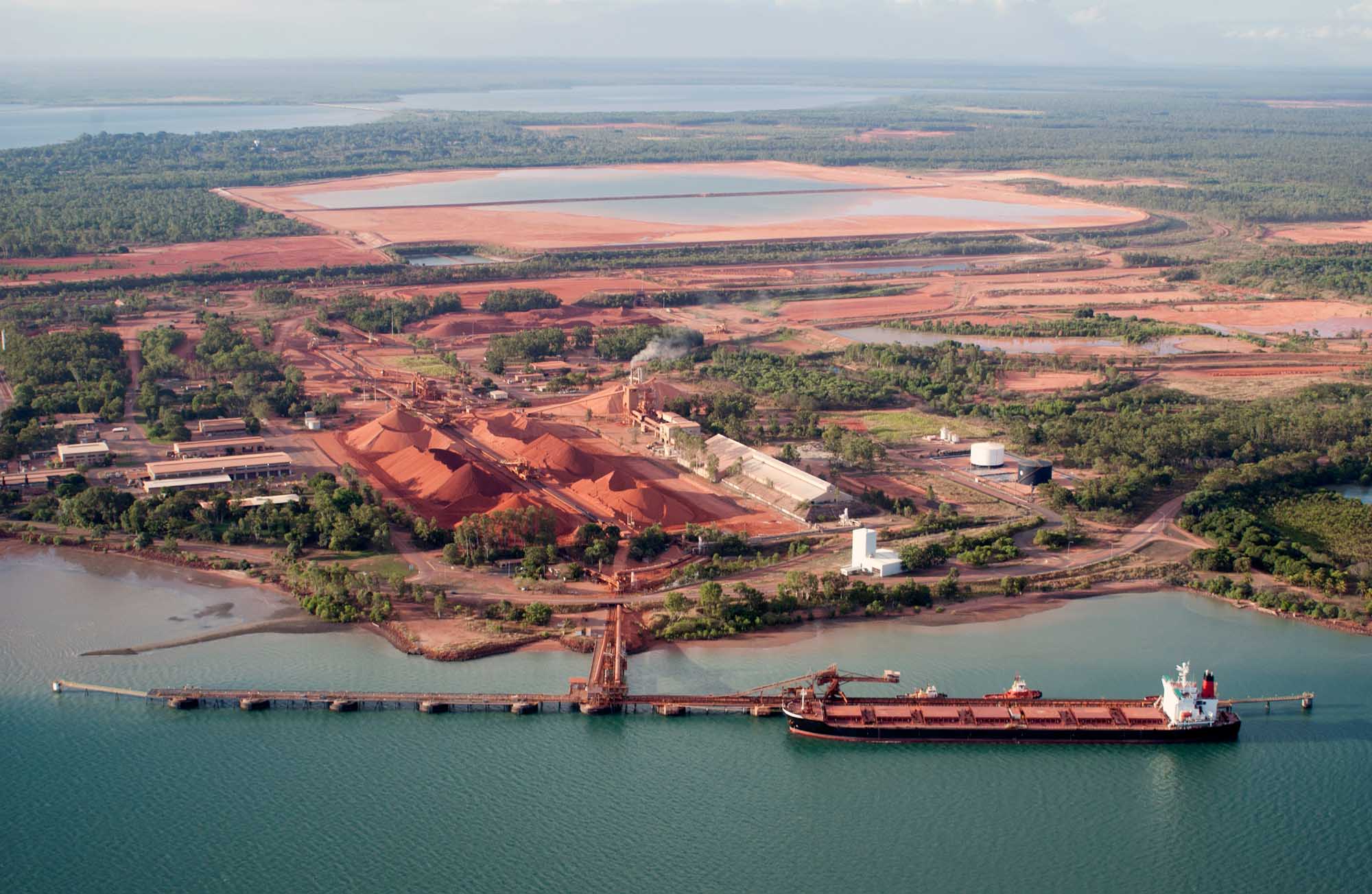 An aerial view of a large-scale bauxite mining and loading operation at the port of Weipa, Queensland, Australia. The image shows massive red ore stockpiles, industrial processing facilities, loading infrastructure, and a cargo vessel docked at the pier, with surrounding cleared mining lands and forested areas visible across the landscape.