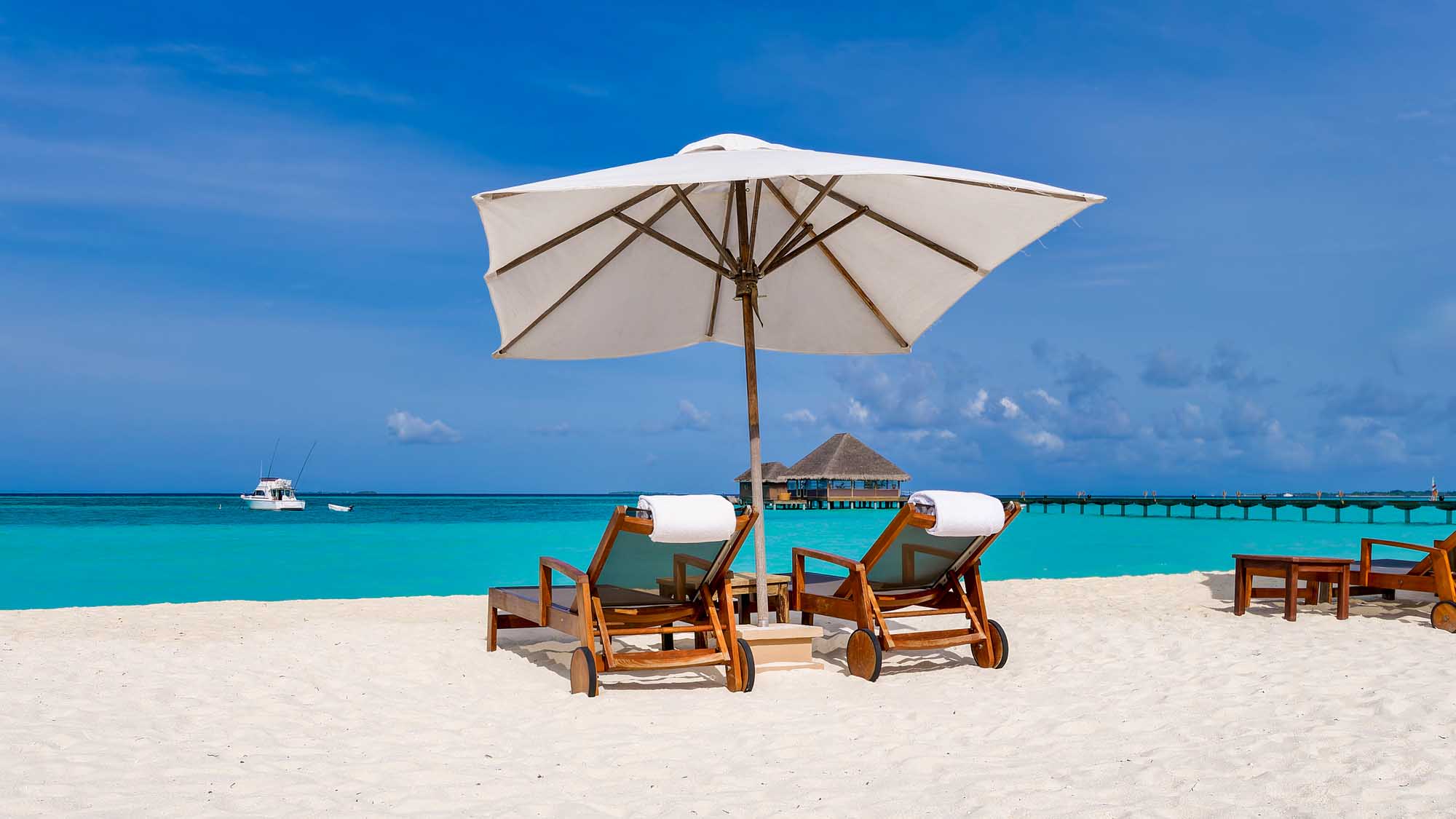 Two wooden sun loungers with white cushions are positioned beneath a large white square umbrella on pristine white sand, overlooking the turquoise waters of the Indian Ocean in the Maldives. A white yacht anchors in the crystal-clear sea, with overwater bungalows and a wooden pier visible in the distance.