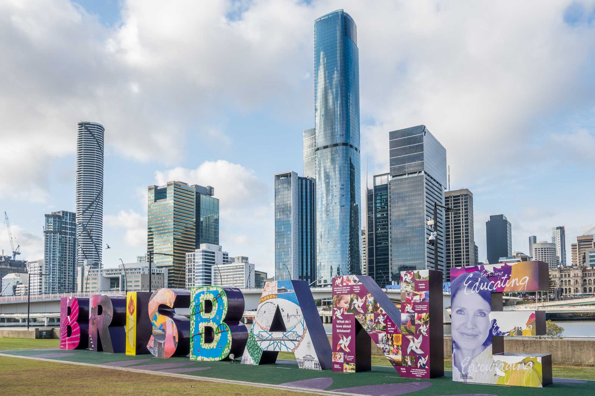 A vibrant, oversized colorful 'BRISBANE' sign installation at South Bank Parklands displays artistic designs and cultural messaging, with the city's modern skyline and skyscrapers rising behind it. The scene captures the iconic landmark against a partly cloudy sky with the Brisbane River visible in the background.
