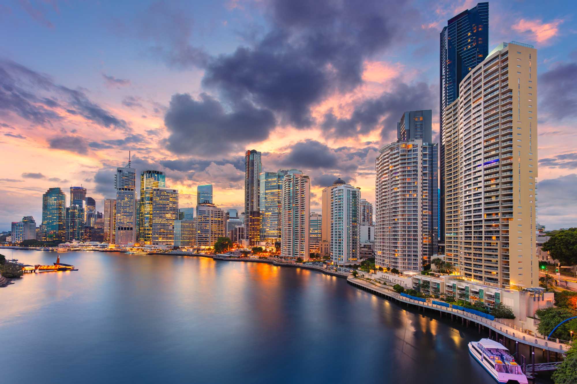 A stunning cityscape of Brisbane, Australia's skyline illuminated during golden hour, featuring modern high-rise buildings reflected in the calm waters of the Brisbane River. The dramatic cloudy sky is painted in warm orange, pink, and purple tones, with passenger ferries visible along the riverbank.