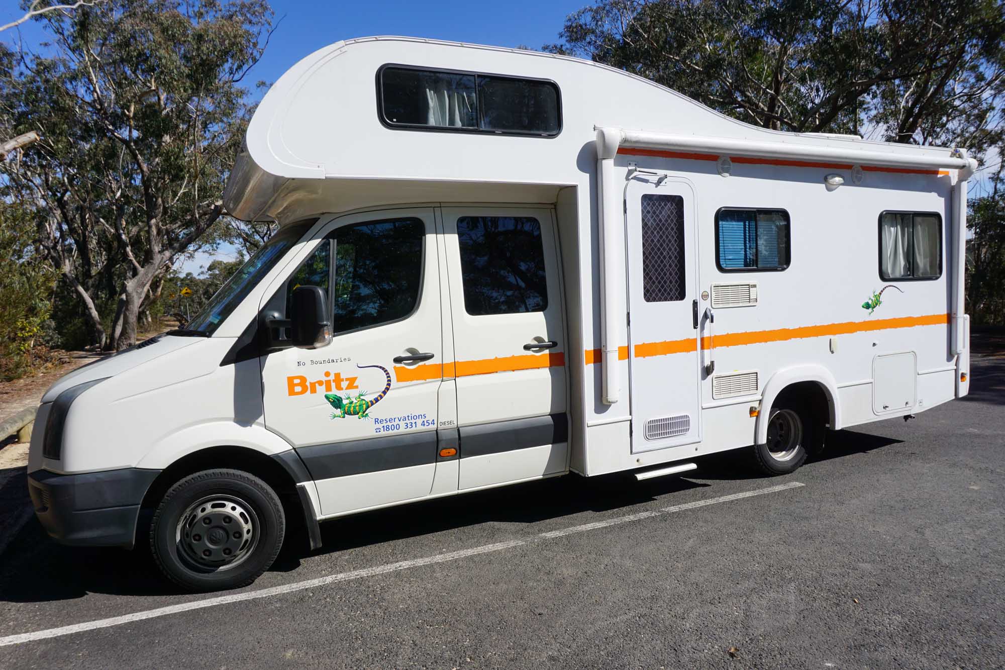 A white and orange Britz campervan is parked on a sunny Australian road lined with eucalyptus trees. Britz is Australia's largest campervan rental company, and this vehicle exemplifies the popular mobile accommodation option for road trips across the country.