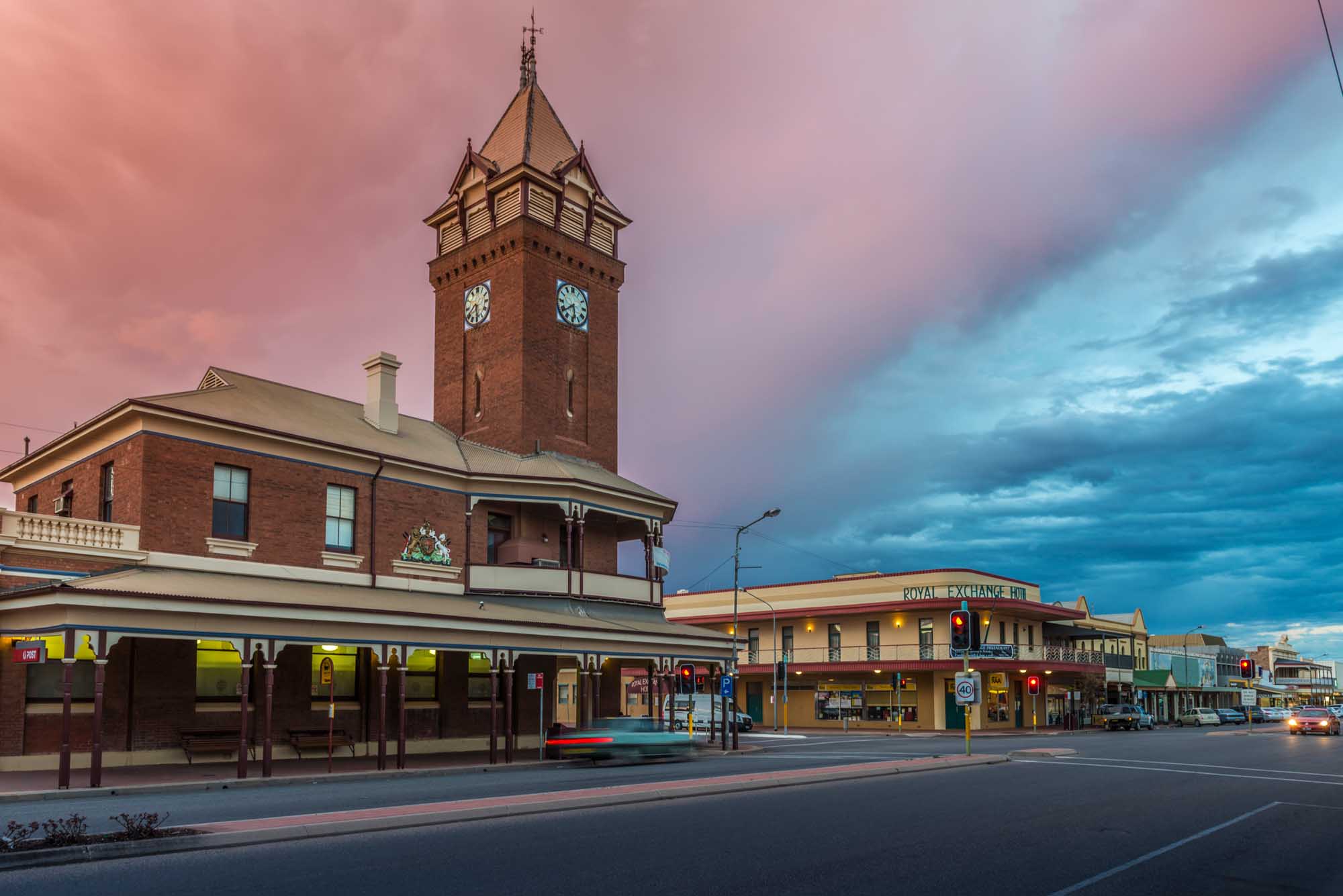 The historic Broken Hill Post Office, built in 1892, stands illuminated against a dramatic twilight sky featuring pink and blue hues. The Victorian-era brick building with its distinctive clock tower is complemented by the Royal Exchange Hotel visible in the background, capturing the architectural heritage of this Australian outback town.