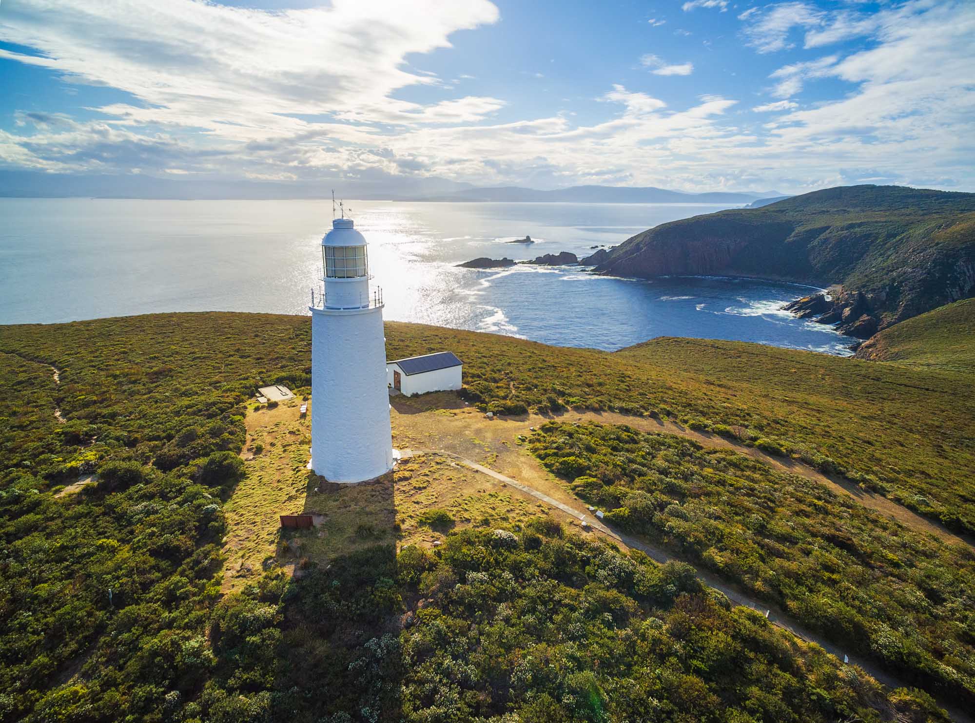 An aerial view of Bruny Island Lighthouse perched on a grassy headland overlooking the Southern Ocean. The white cylindrical lighthouse stands prominently on the rugged coastline with dramatic cliffs, rocky outcrops, and dense coastal vegetation, with sparkling blue waters and distant landmasses visible under a partly cloudy sky.