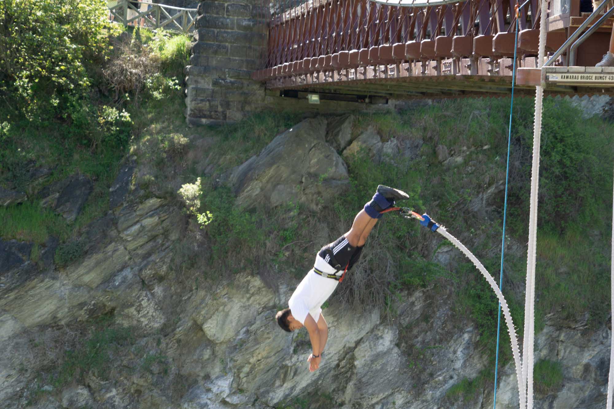 A thrill-seeker mid-jump during a bungy leap from the iconic Kawarau Bridge near Queenstown, New Zealand, suspended above the river gorge with a safety rope attached. The dramatic rocky canyon walls and lush vegetation frame this adrenaline-pumping adventure activity.