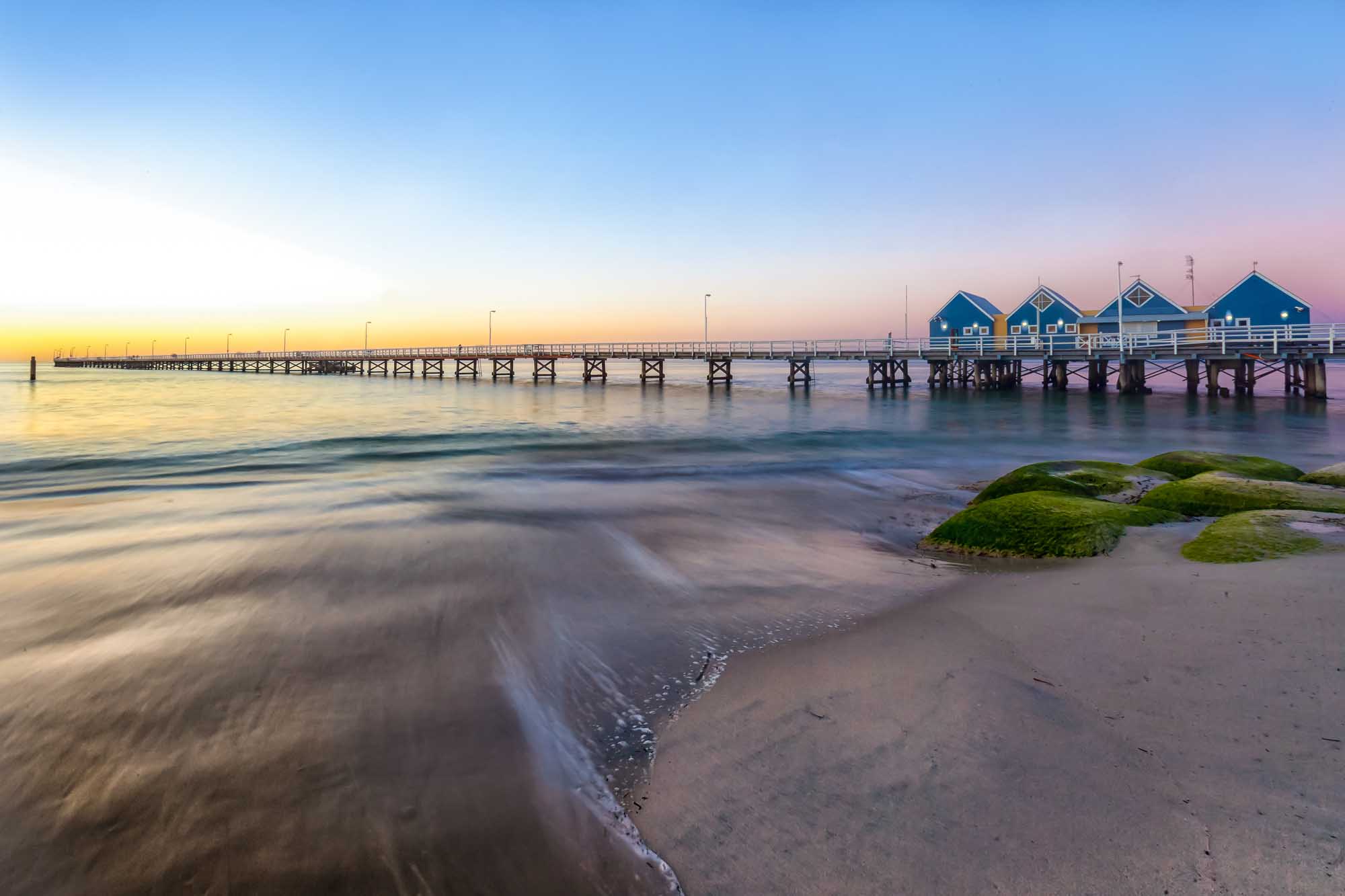 A serene coastal scene of the iconic Busselton Jetty stretching into calm waters at dawn, with distinctive blue boathouses visible at the end of the pier. Moss-covered rocks dot the foreground sandy beach, while soft golden and purple hues paint the sky during the early morning light.