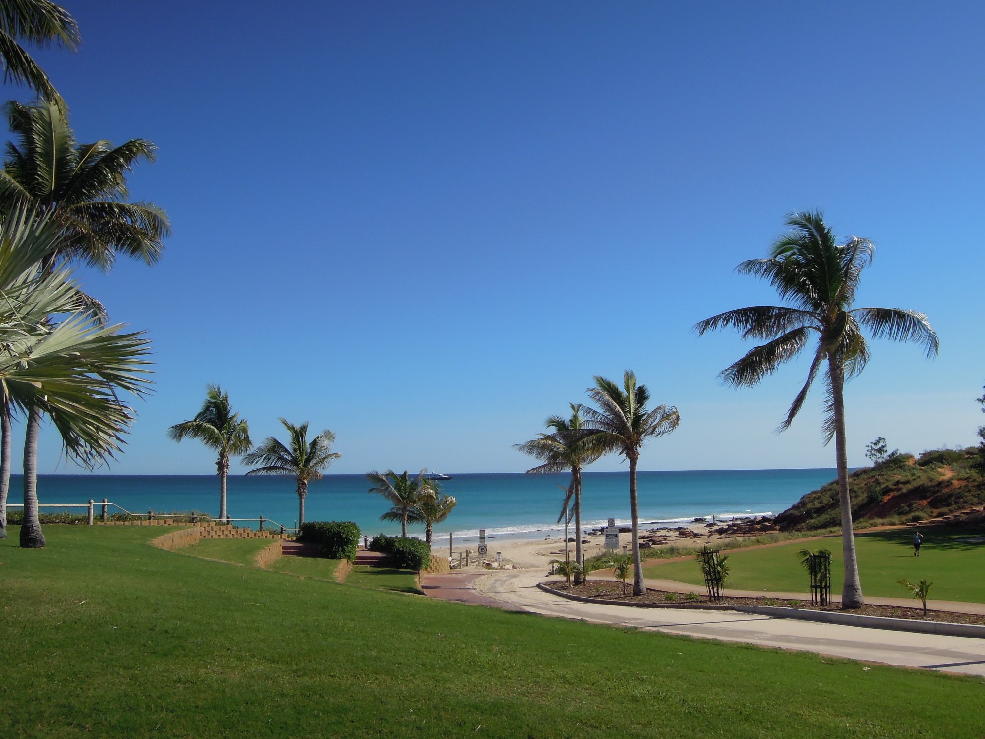 A scenic beachfront park with manicured lawns, palm trees, and clear ocean views overlooking Cable Beach in Broome. The image captures a serene coastal destination with pristine turquoise waters, sandy beach, and well-maintained recreational grounds perfect for visitors.