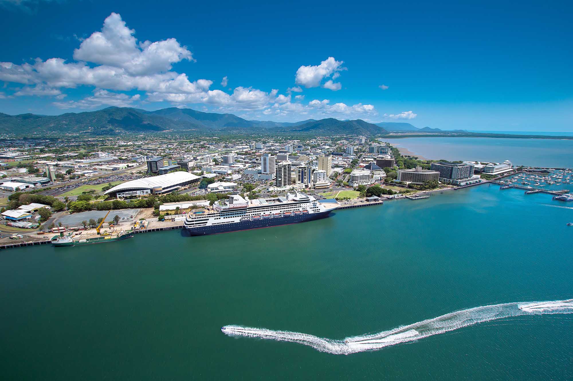 An aerial view of Cairns, Queensland, Australia, showcasing the vibrant coastal city with a large cruise ship docked at the port, surrounded by turquoise waters, sandy beaches, and tropical mountains in the background under a bright blue sky with white clouds.