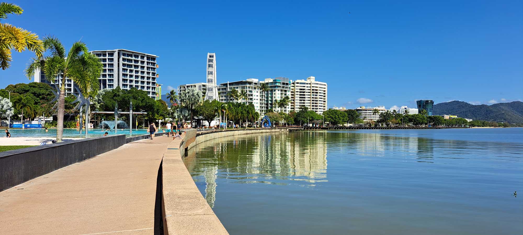 A scenic waterfront promenade in Cairns, Australia, featuring a calm lagoon, palm-lined esplanade with public pool facilities, and modern high-rise residential and commercial buildings reflected in the still water under a clear blue sky.