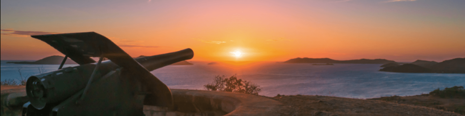 A silhouetted cannon mounted on a coastal fortification overlooks a tranquil bay at golden hour, with distant islands visible across the water and a vibrant sunset sky painting the horizon in warm orange and pink tones.