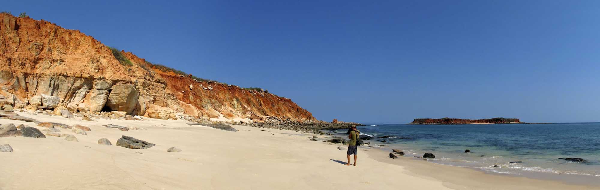 A scenic coastal landscape at Cape Leveque featuring distinctive red and ochre sandstone cliffs overlooking a pristine sandy beach with turquoise waters. A solitary beachgoer stands on the white sand with rocky outcrops and a small island visible in the distance under clear blue skies.
