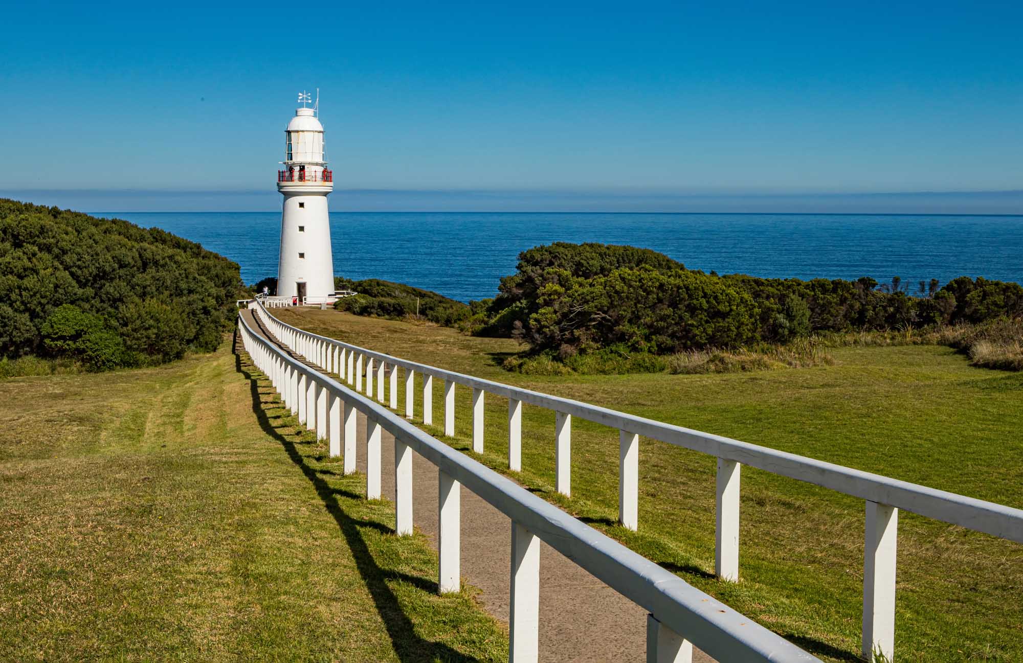A picturesque white lighthouse with red-brown accents stands on a grassy headland overlooking the turquoise ocean, framed by native Australian coastal vegetation. A white safety railing leads toward the lighthouse, exemplifying the scenic beauty of Australia's Great Ocean Road.