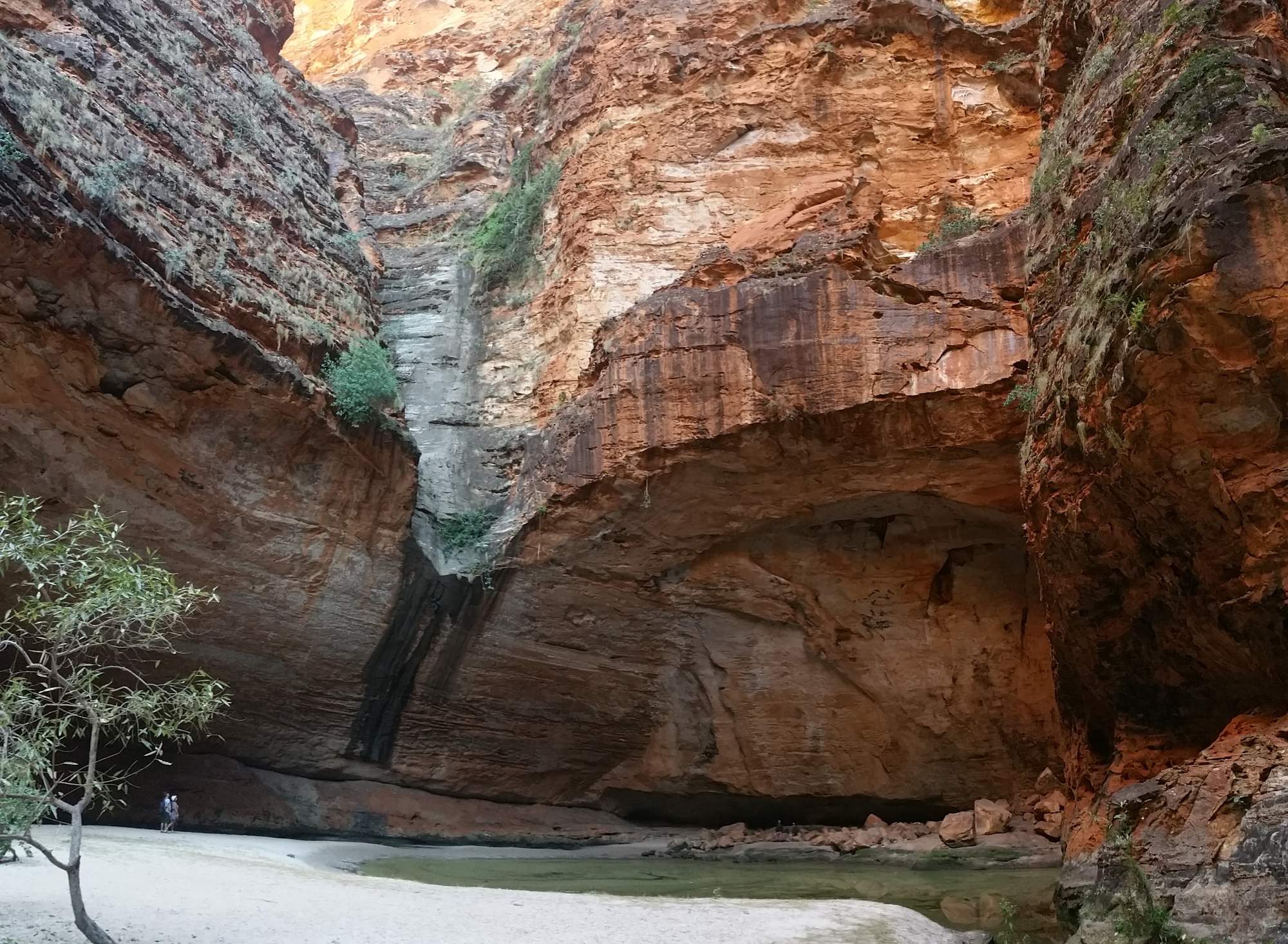 A dramatic canyon landscape featuring towering stratified rock walls flanking a narrow gorge with a small waterfall and pool of water. Lush green vegetation clings to the cliff faces, while visitors explore the sandy canyon floor beneath the imposing natural formations.