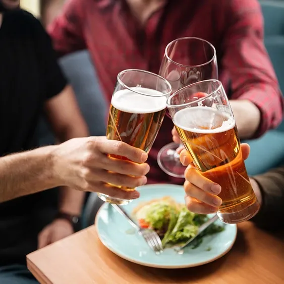 Three passengers clink glasses of beer at the Empress Bar aboard the Cordelia cruise ship, with a fresh salad plate visible on the table. The scene captures a convivial moment during a casual meal or social gathering onboard.