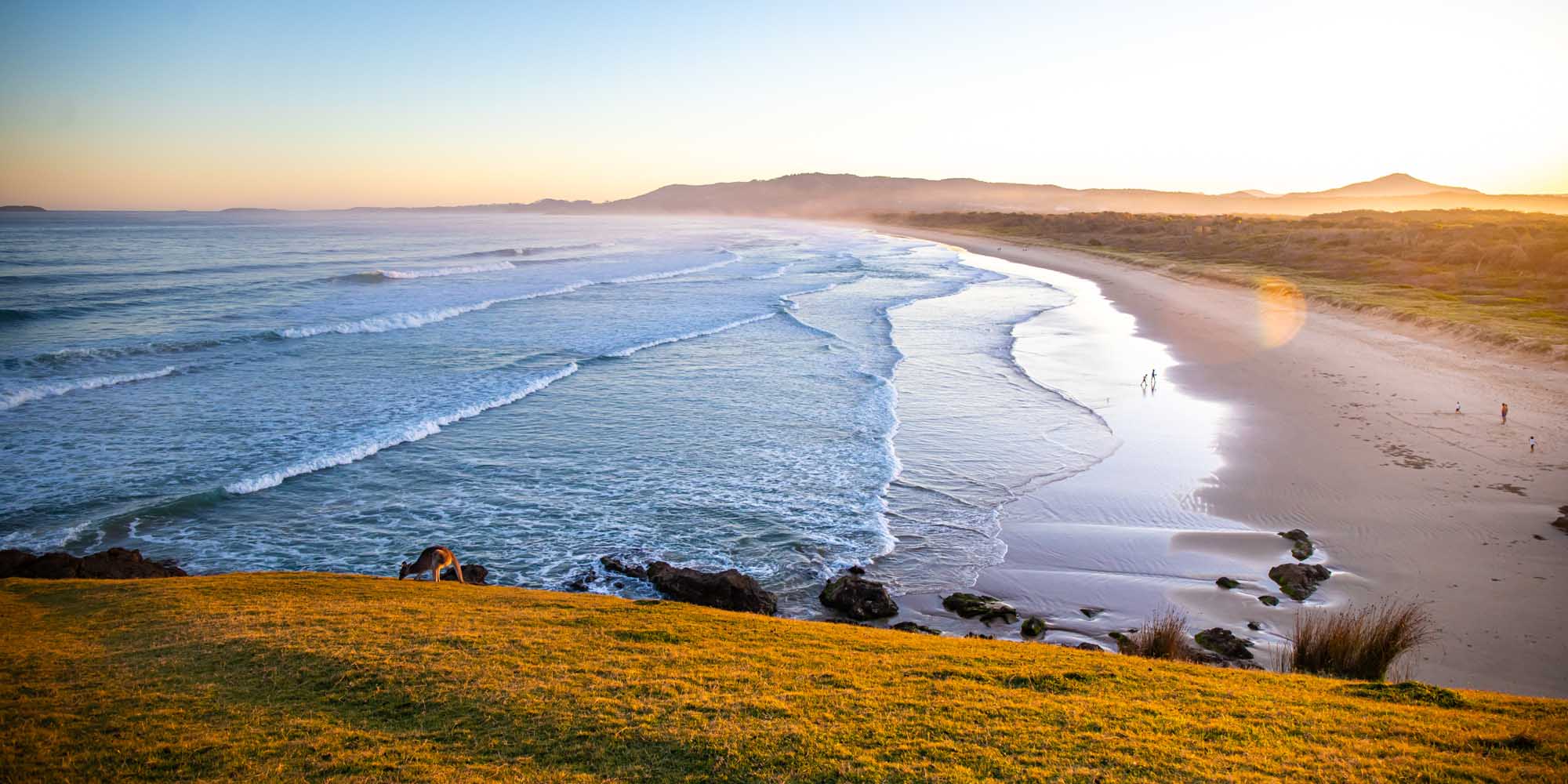 A stunning coastal landscape featuring a pristine Australian beach at golden hour, with rolling waves, sandy shores, and rolling hills in the background. The scene captures the natural beauty of New South Wales with its characteristic coastline, sparse vegetation, and dramatic lighting as the sun sets over the ocean.