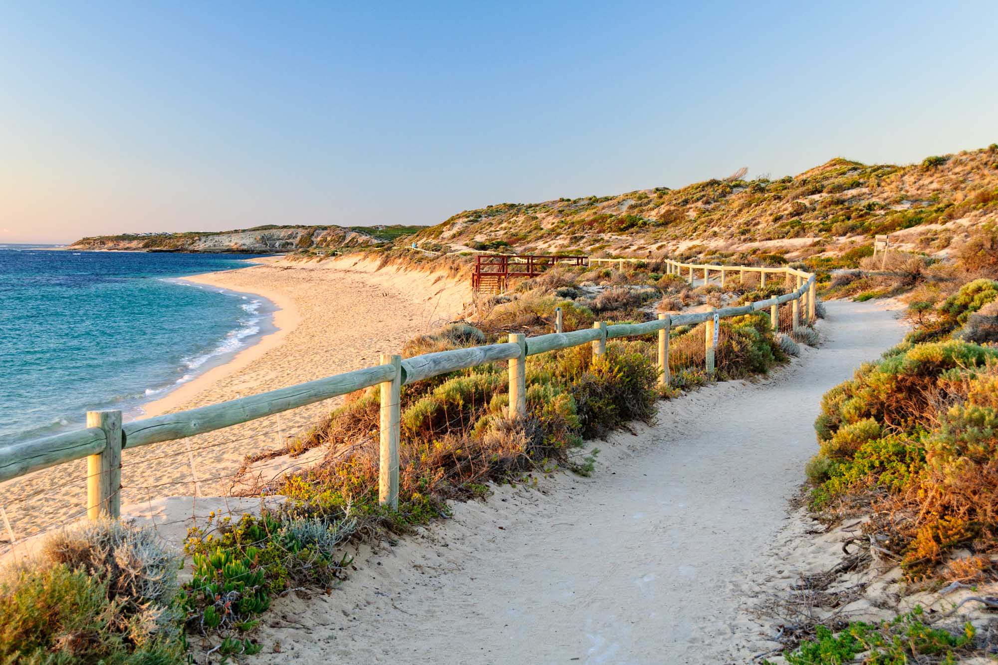 A scenic wooden-railed walking path winds along the pristine sandy shores of Gnarabup Beach near Prevelly, Western Australia. The path overlooks turquoise waters, golden sand, and native coastal vegetation clinging to the dramatic ochre cliffs, with a red lifeguard tower visible in the distance.