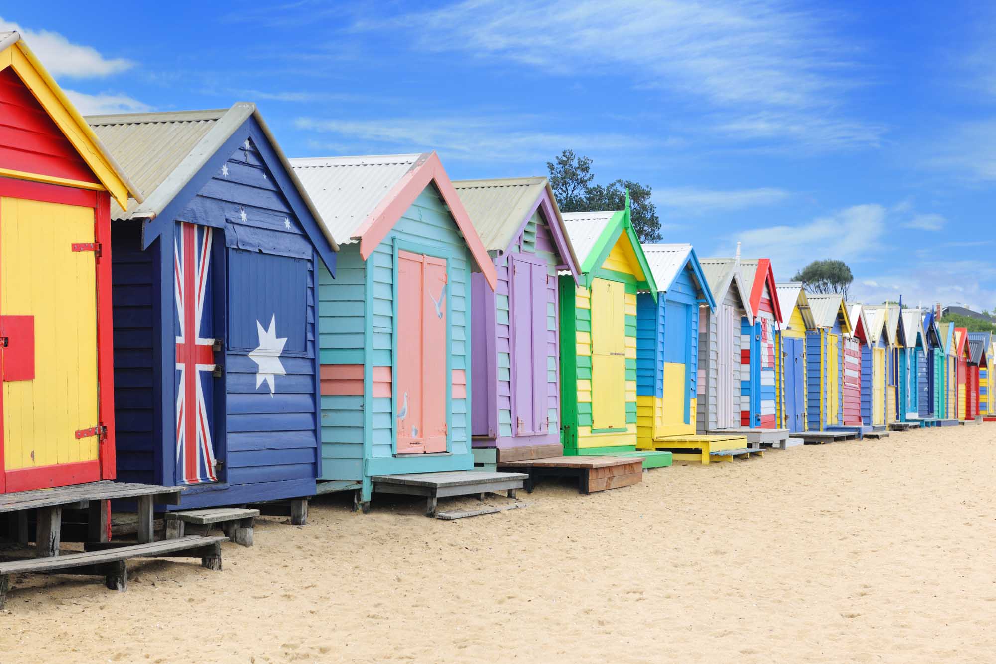 A vibrant row of traditionally-styled beach huts in bright primary colors lines the sandy shore at Brighton Beach in Melbourne, Australia. These iconic changing rooms are among the most expensive real estate per square foot in the country and represent a beloved Australian coastal heritage landmark.