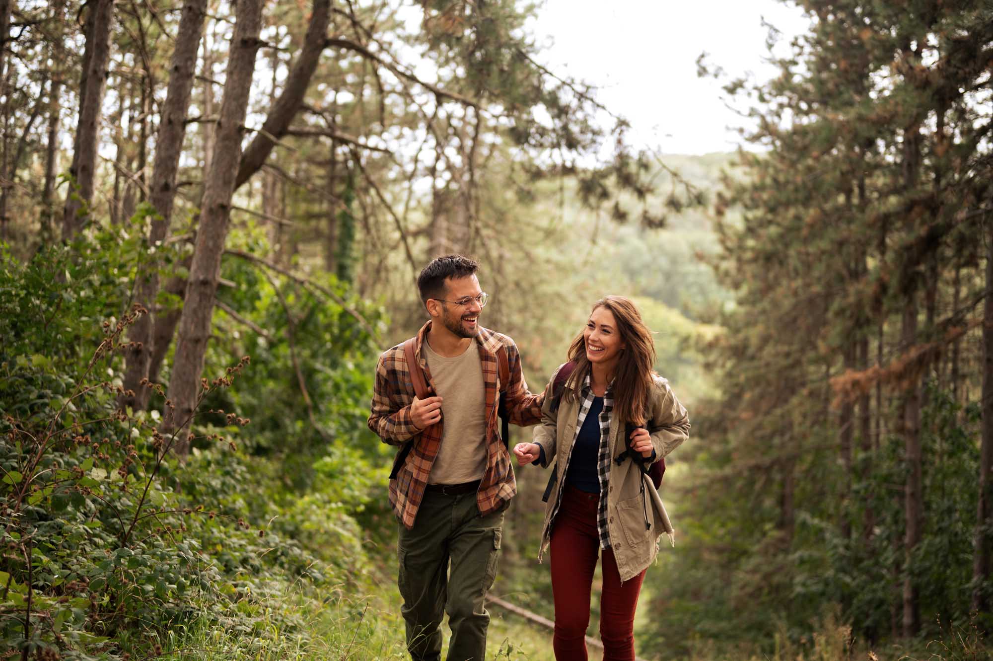 A young couple jogging together on a forest trail surrounded by tall evergreen trees and lush green vegetation. Both are smiling and engaged in conversation as they explore the natural woodland landscape.