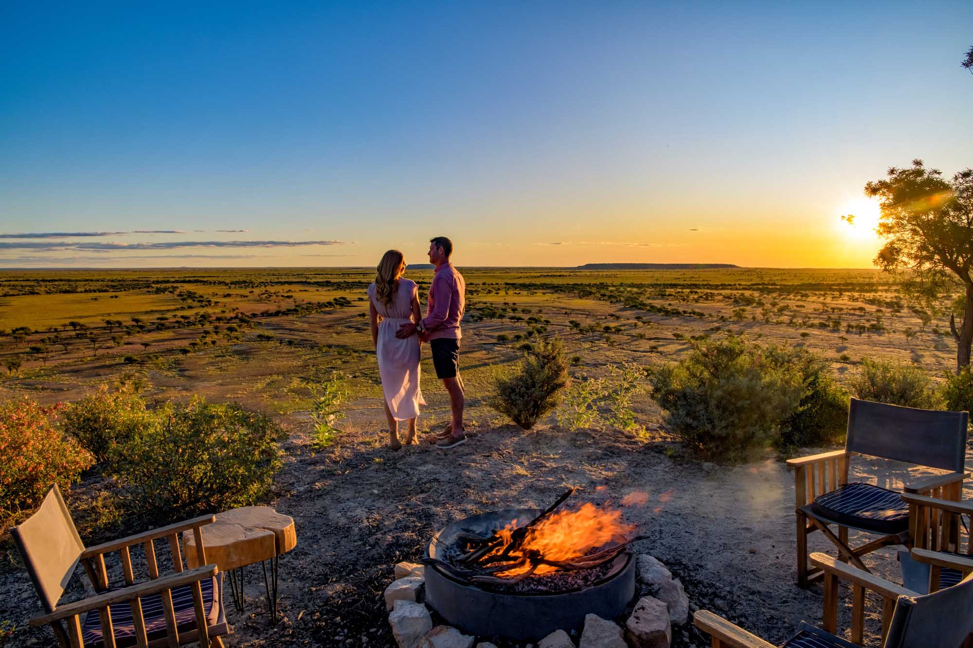 A couple stands together overlooking a vast, arid Australian landscape dotted with sparse vegetation at golden hour, with a campfire burning in a metal fire pit in the foreground. Empty wooden chairs flank the fire, creating an intimate outdoor camping or glamping setting in the remote Outback.