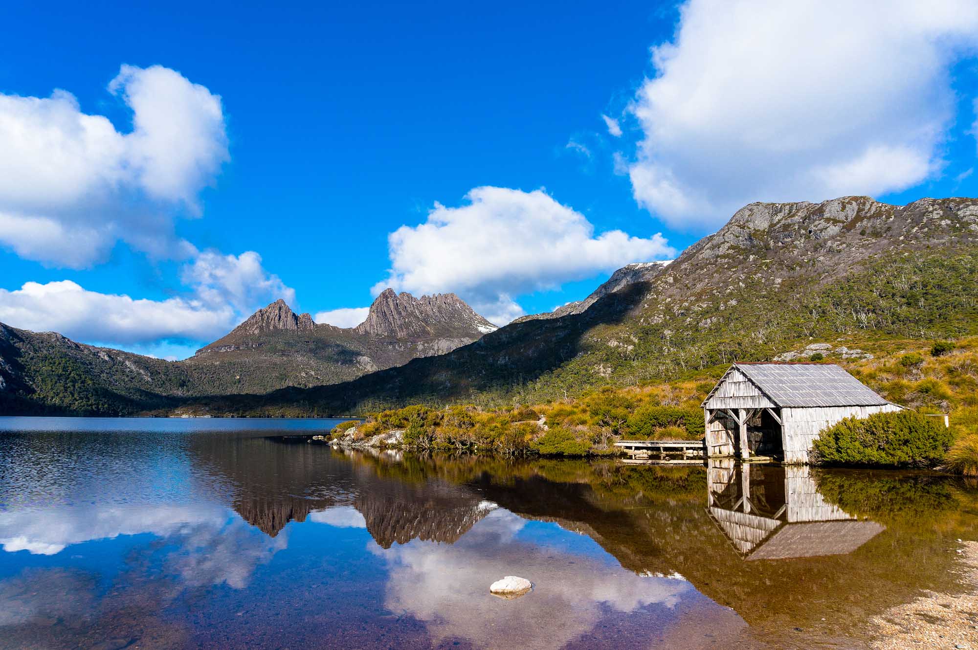 A picturesque alpine landscape featuring the iconic jagged peaks of Cradle Mountain reflected in the still waters of Dove Lake, with a weathered wooden boat shed nestled along the shoreline surrounded by native vegetation. This UNESCO World Heritage site in Tasmania showcases pristine natural beauty under a brilliant blue sky with white clouds.