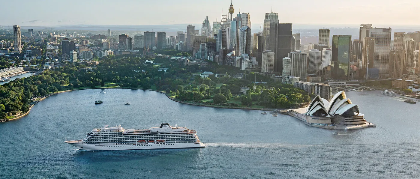 A large modern cruise ship navigates Sydney Harbour with the iconic Sydney Opera House prominently featured on the right, the Sydney skyline in the background, and lush green parkland along the waterfront. The scene captures one of the world's most recognizable harbour destinations during daytime with clear visibility.
