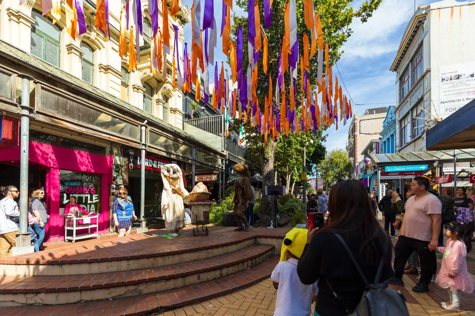 A vibrant street celebration in Wellington, New Zealand featuring colorful purple and orange ribbon decorations strung overhead, street performers in costume, and crowds of visitors enjoying the festive Cuba Dupa Festival atmosphere in an urban pedestrian area lined with shops and historic buildings.