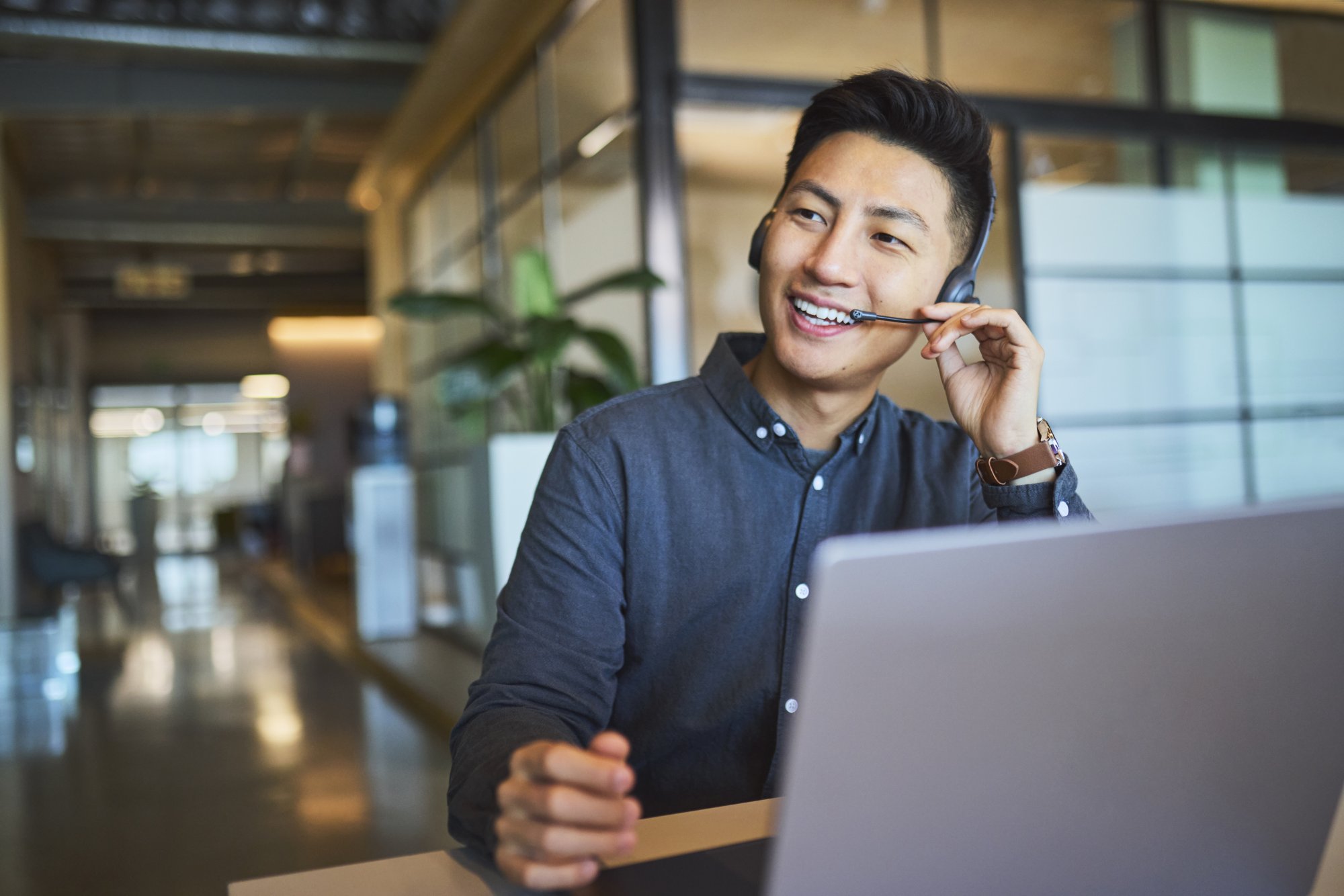 A young professional wearing a headset and dark button-up shirt sits at a laptop in a modern office