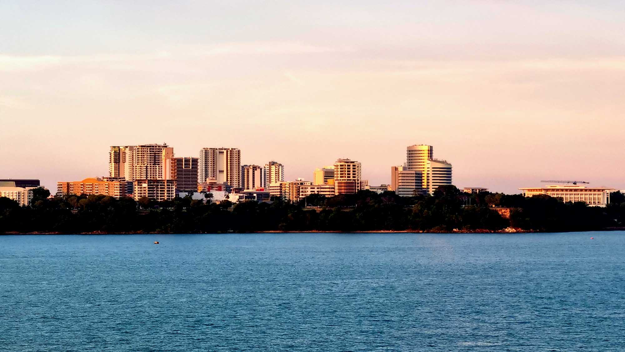 A scenic waterfront view of Darwin's modern skyline illuminated by golden sunset light, with multi-story residential and commercial buildings rising above lush tropical vegetation along the harbor. The calm blue waters reflect the warm glow of the late afternoon sun across the cityscape.