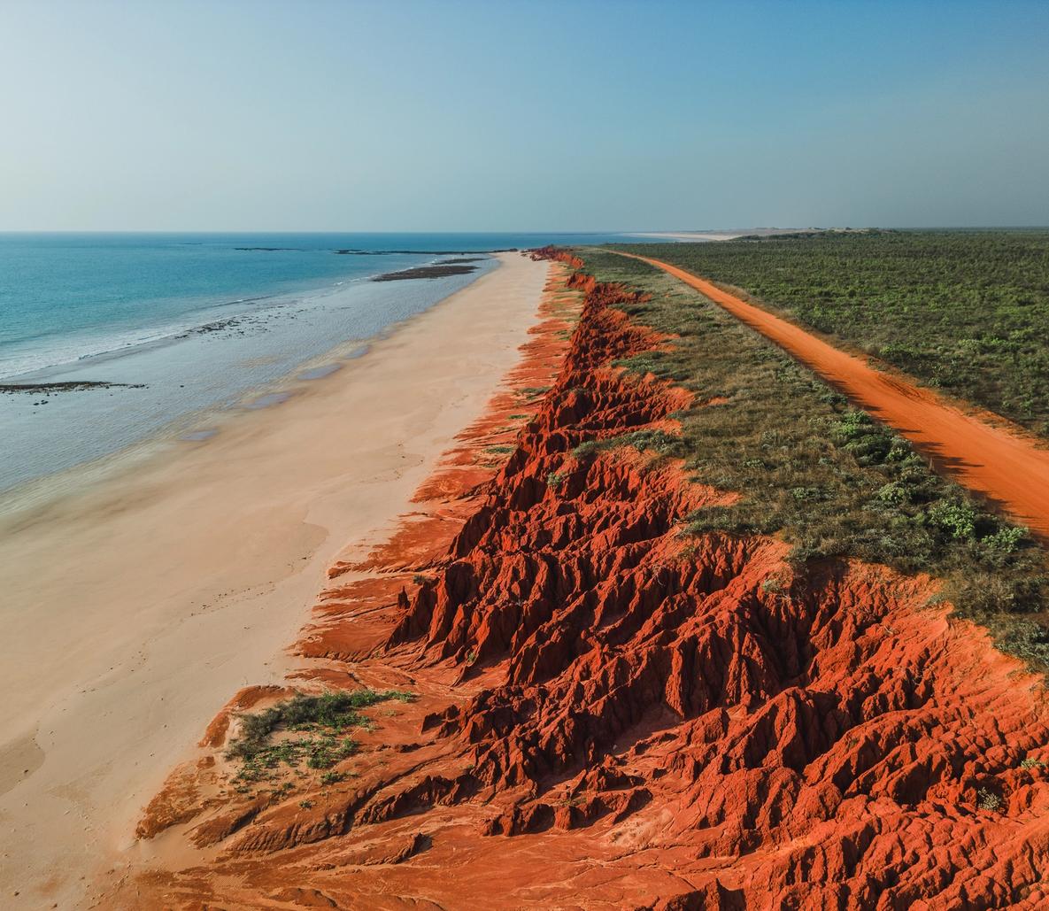 A stunning aerial view of a striking coastline where vibrant red-orange cliffs with deep erosion