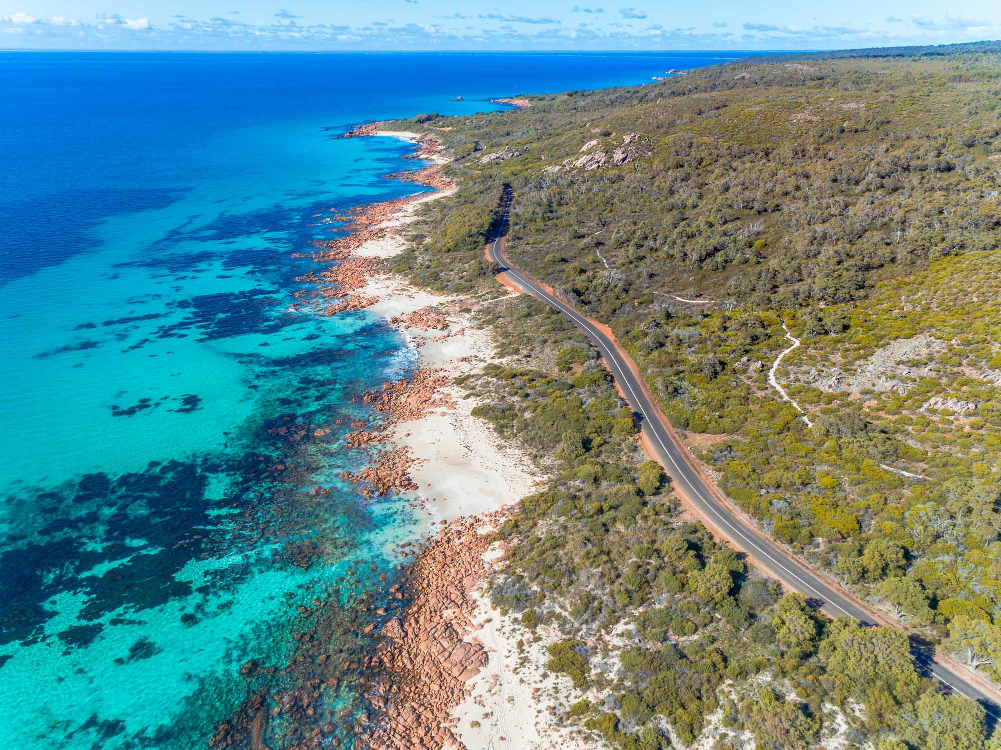 An aerial view of a scenic coastal road hugging a pristine sandy beach with turquoise waters and red rocky outcrops along the shoreline. The road winds through native bushland under bright summer sunlight, showcasing the natural beauty of Western Australia's coastline.