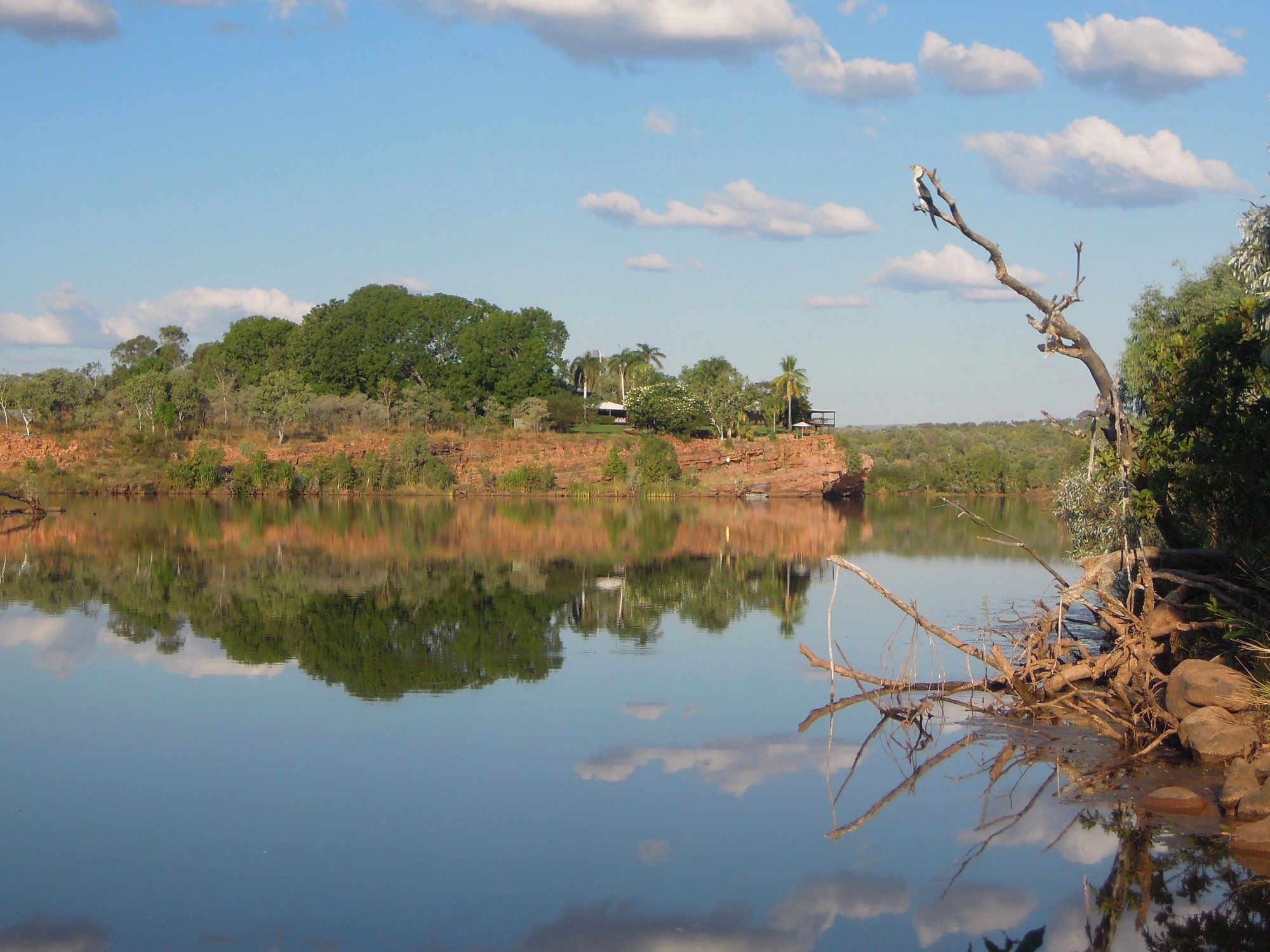 A serene riverbank view at El Questro Station in Western Australia, featuring calm reflective waters, red soil cliffs, native vegetation, and scattered buildings nestled in the savanna landscape under a partly cloudy sky.